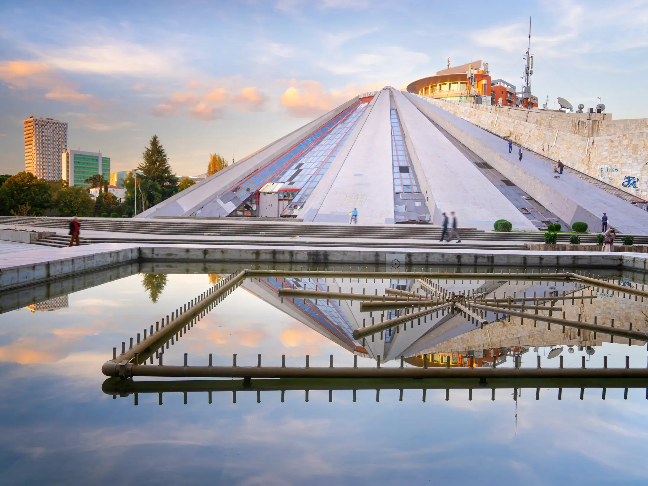 Die sogenannte Pyramide von Tirana in Albanien spiegelt sich im Abendlicht in einem Wasserbecken vor der Skyline der Stadt.