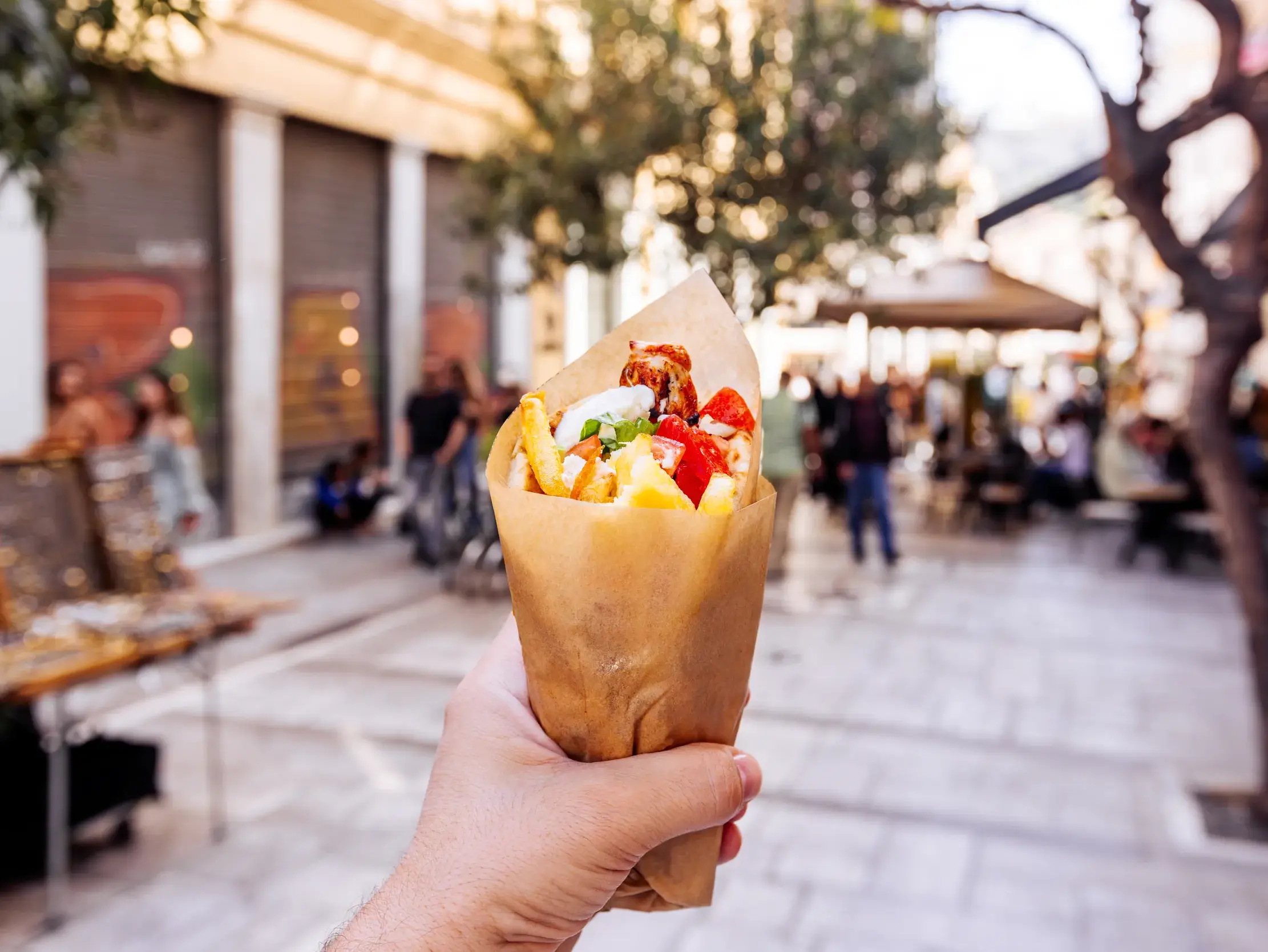 Eine Hand hält eine frische Gyros-Pita mit Pommes in einer Papiertüte vor der Kulisse einer belebten Einkaufsstraße in Athen.