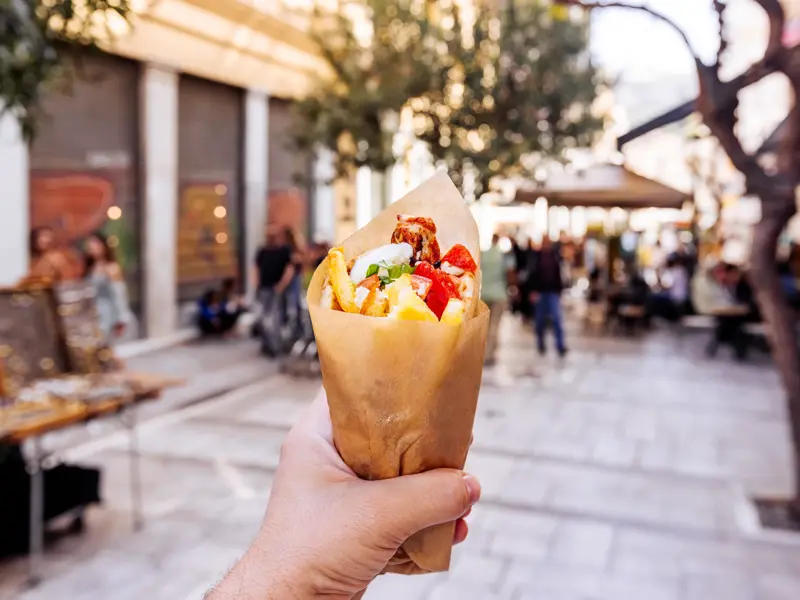 Eine Hand hält eine frische Gyros-Pita mit Pommes in einer Papiertüte vor der Kulisse einer belebten Einkaufsstraße in Athen.