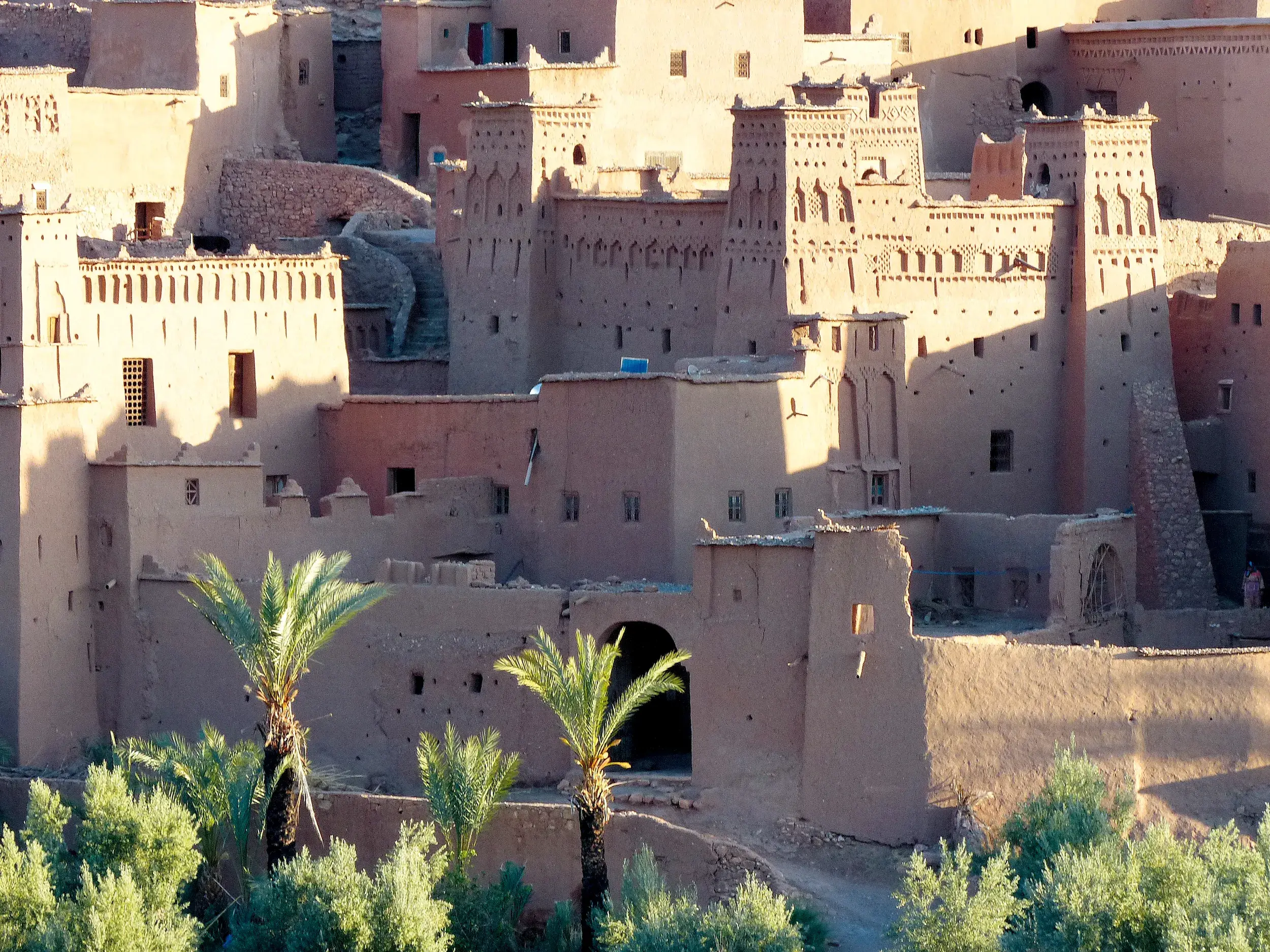 Historische Lehmbauten des Ksar Ait Benhaddou, UNESCO-Weltkulturerbe in Marokko, ragen hinter grünen Palmen empor.