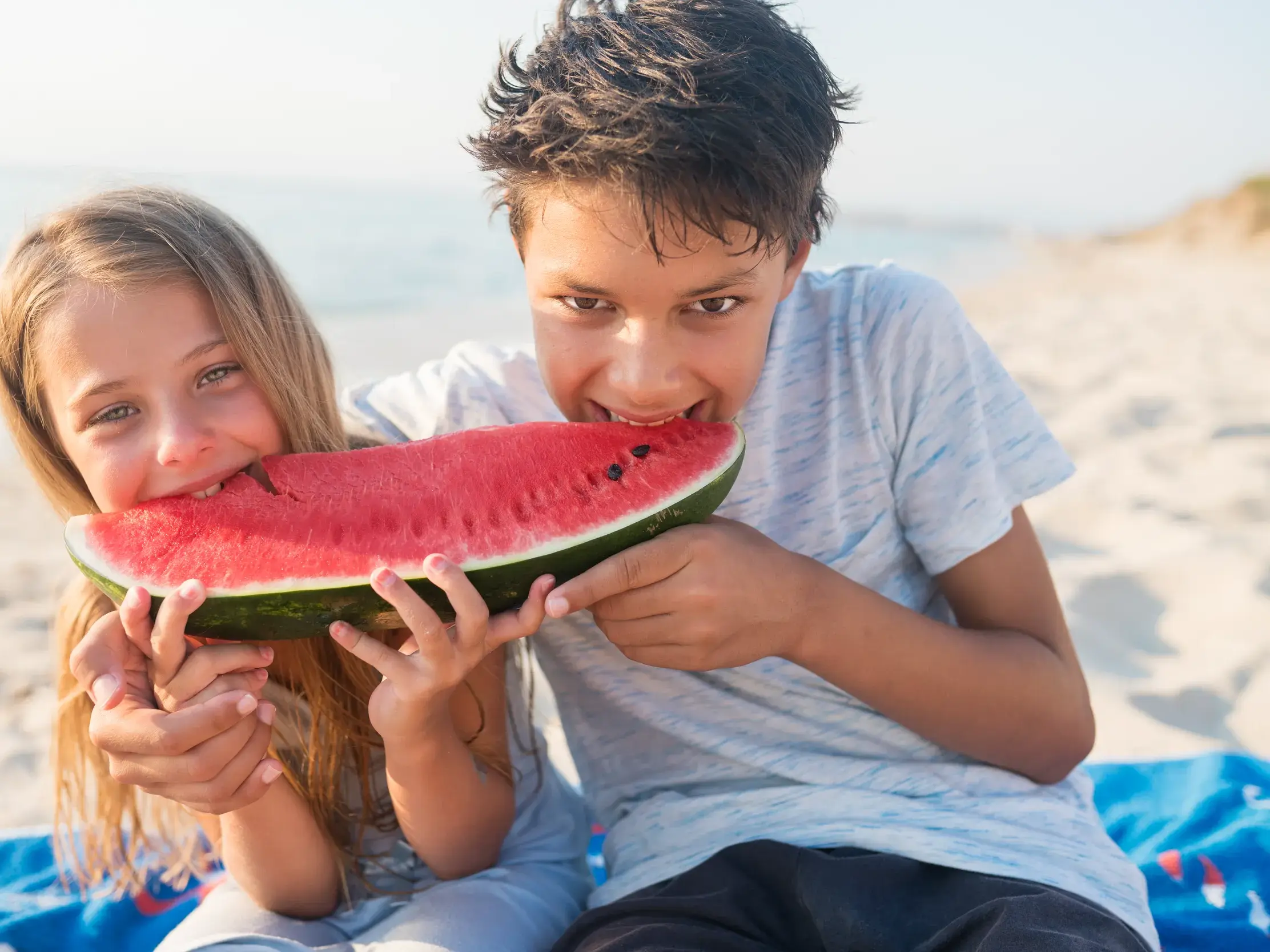 Zwei Kinder genießen lachend ein großes Stück Wassermelone am Sandstrand während ihres Familienurlaubs in Griechenland.