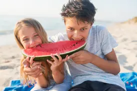 Zwei Kinder genießen lachend ein großes Stück Wassermelone am Sandstrand während ihres Familienurlaubs in Griechenland.