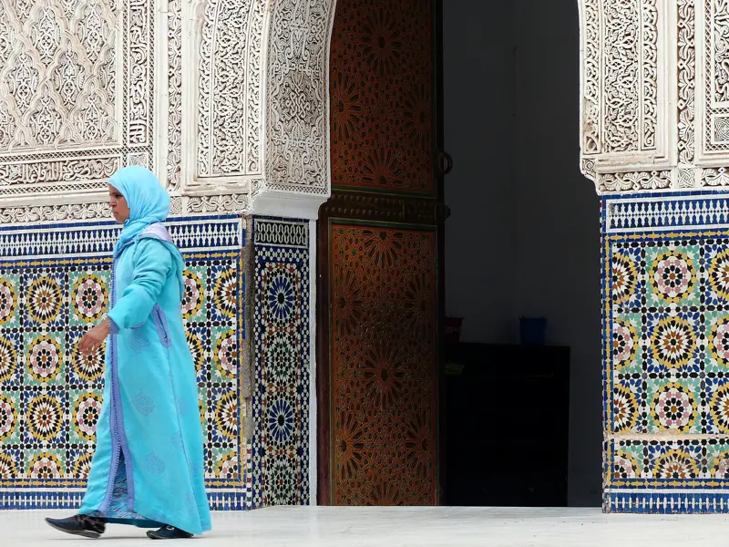 Eine Frau in hellblauer Djellaba geht an einer prachtvollen Wand mit Mosaiken und Stuckverzierungen in Marrakesch vorbei.