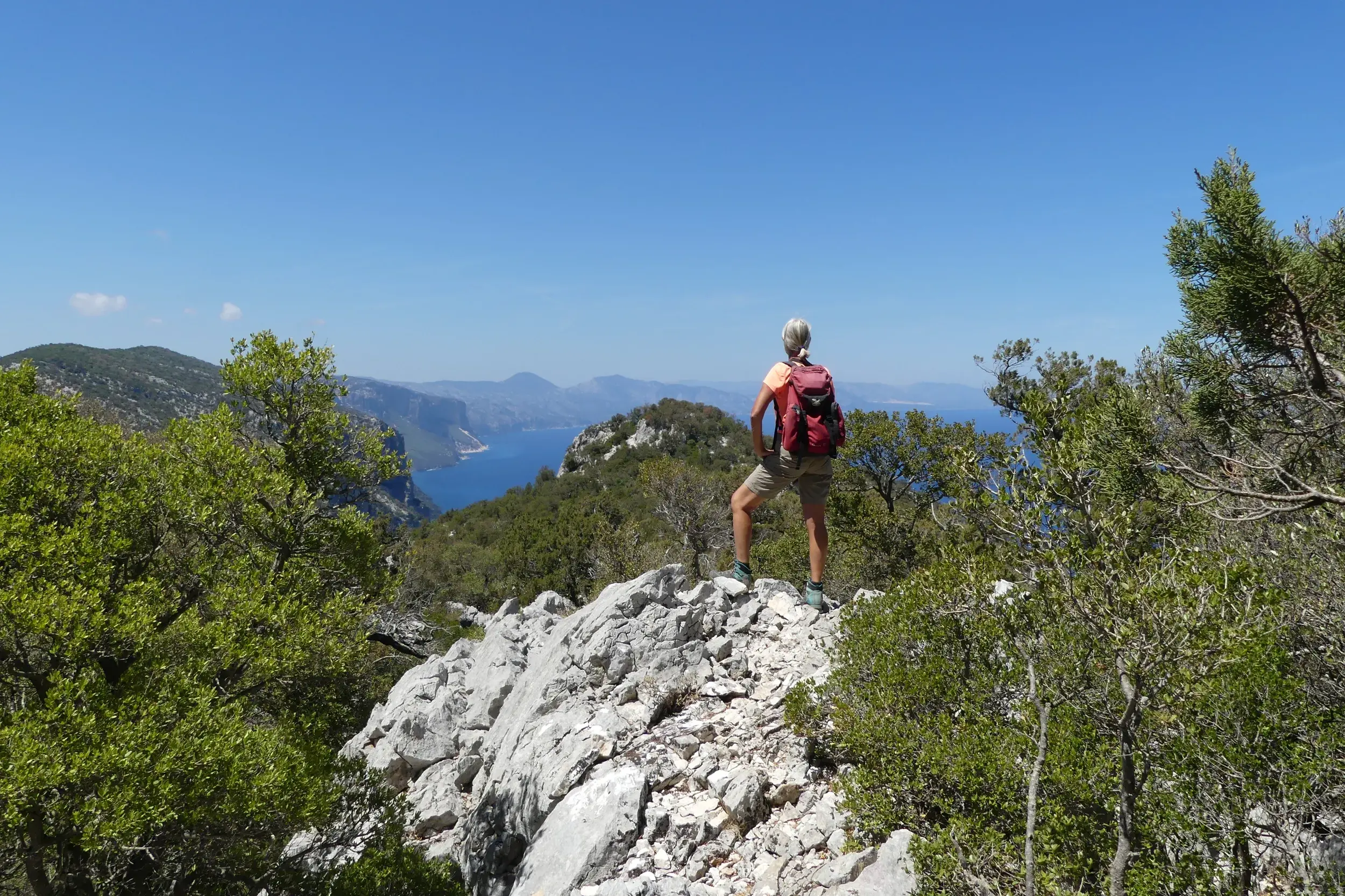 Wanderin mit Rucksack blickt von einem Felsvorsprung im Supramonte über die Küste des Golfs von Orosei auf Sardinien.
