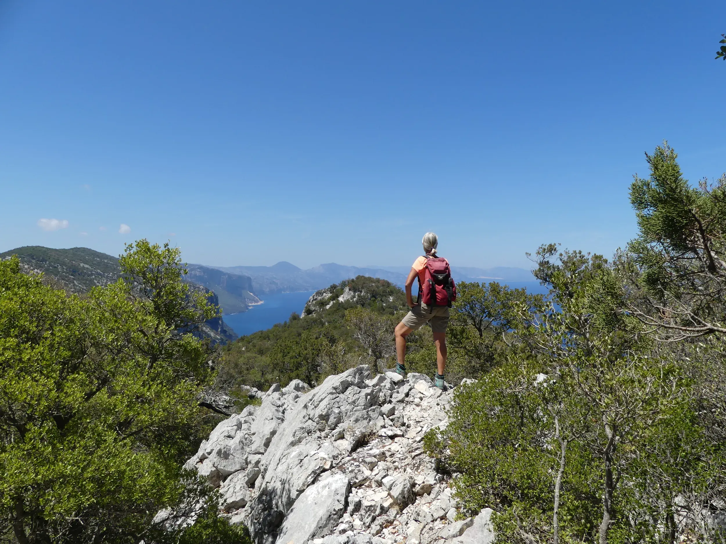 Wanderin mit Rucksack blickt von einem Felsvorsprung im Supramonte über die Küste des Golfs von Orosei auf Sardinien.