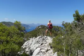 Wanderin mit Rucksack blickt von einem Felsvorsprung im Supramonte über die Küste des Golfs von Orosei auf Sardinien.
