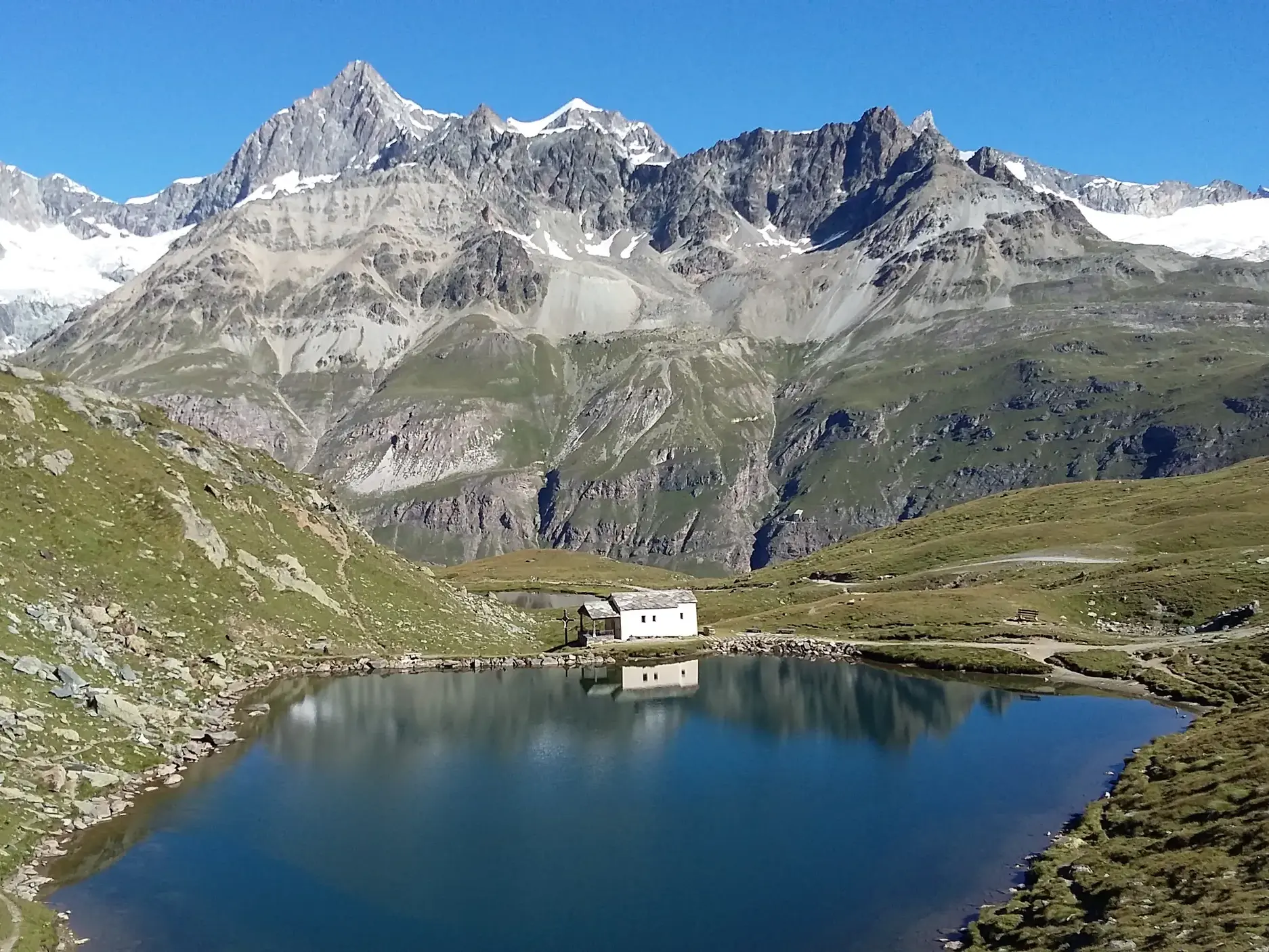 Kleine Hütte an einem klaren Bergsee, in dem sich die schneebedeckten Gipfel der Walliser Bergwelt bei einer Wanderung spiegeln.