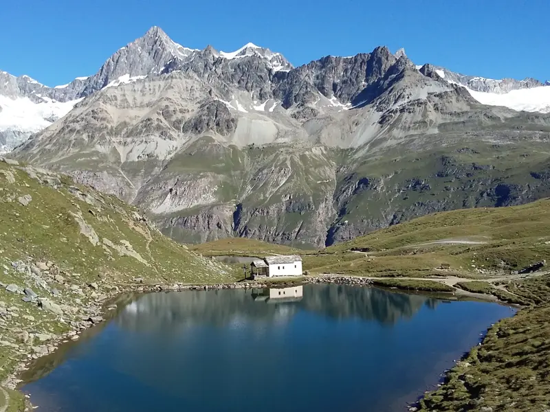Kleine Hütte an einem klaren Bergsee, in dem sich die schneebedeckten Gipfel der Walliser Bergwelt bei einer Wanderung spiegeln.