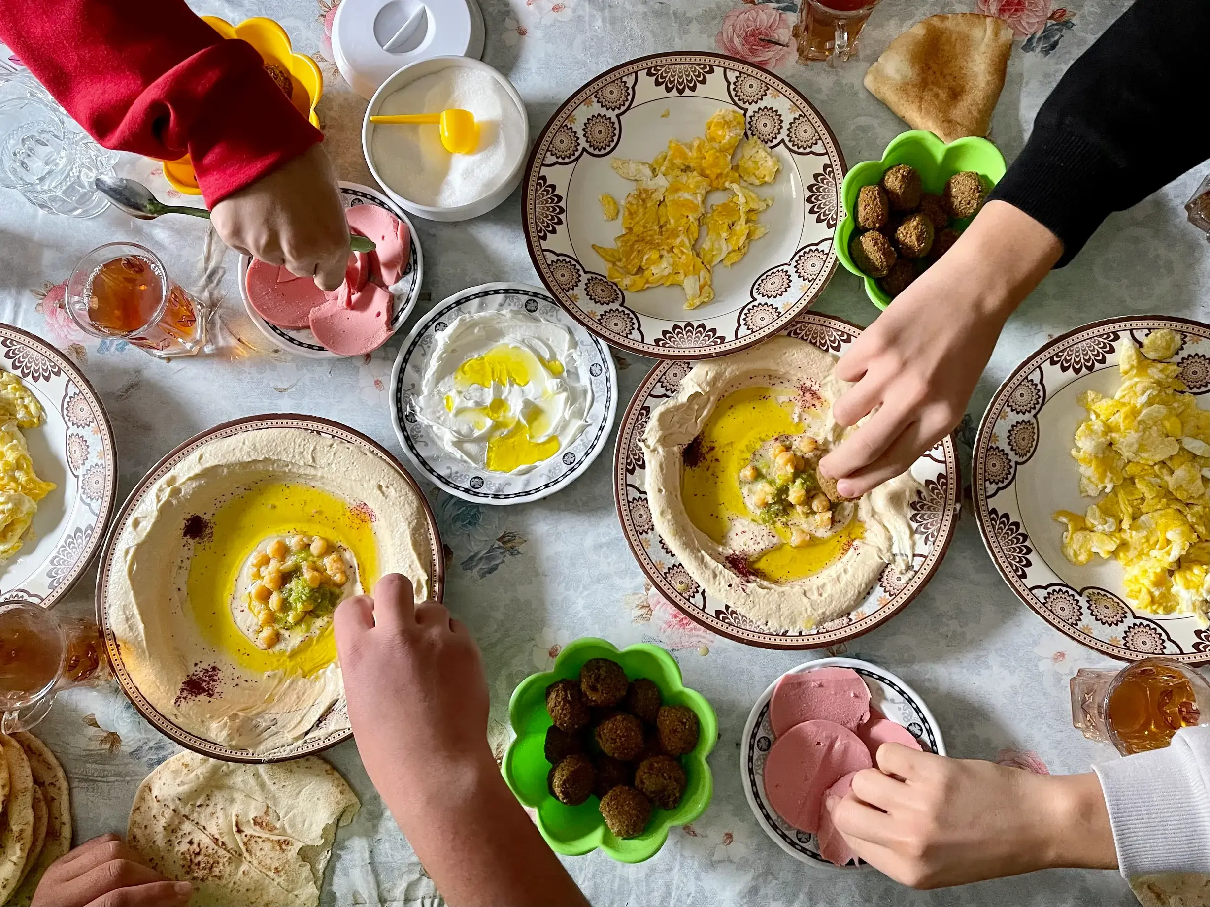 Hände einer kleinen Gruppe greifen nach Hummus, Falafel und Rührei auf einem reich gedeckten Frühstückstisch in Jordanien.