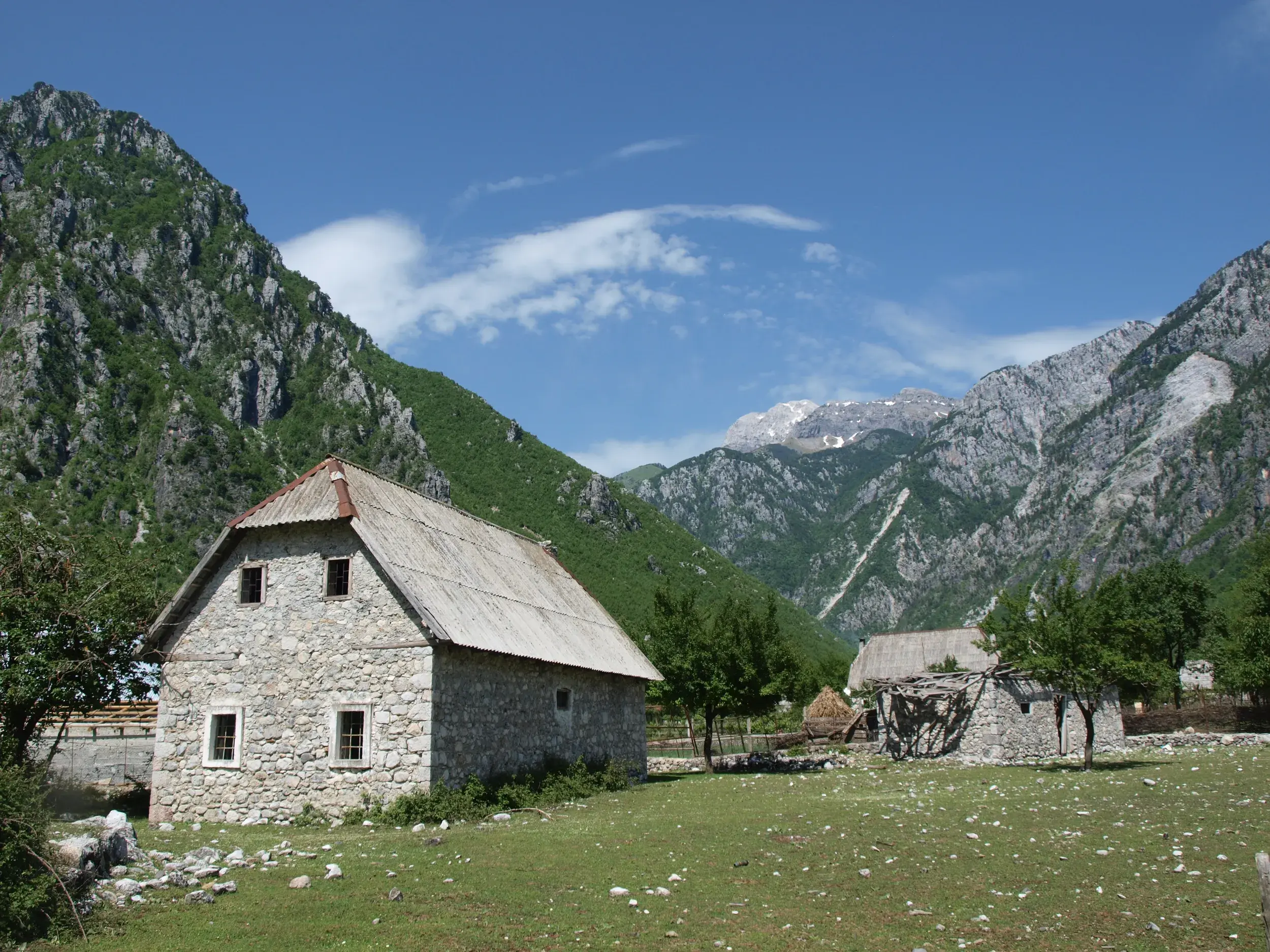 Ein traditionelles Steinhaus auf einer grünen Wiese im Valbona-Tal, umgeben von den beeindruckenden Gipfeln der Albanischen Alpen.