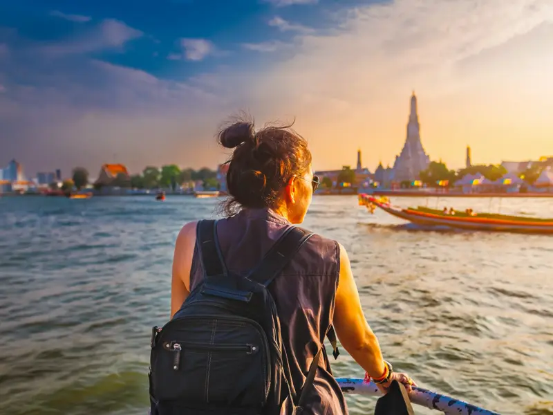 Alleinreisende mit Rucksack blickt bei Sonnenuntergang vom Boot auf den Tempel Wat Arun am Chao Phraya Fluss in Bangkok.
