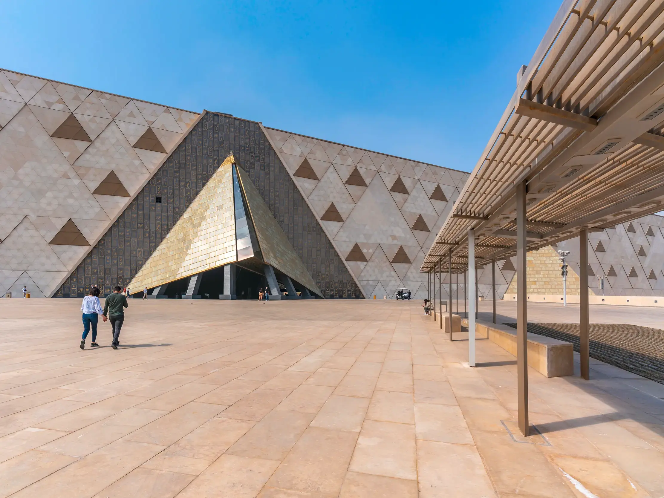 Ein Paar geht an einem sonnigen Tag über den großen Vorplatz zum pyramidenförmigen Eingang des Grand Egyptian Museum in Gizeh.