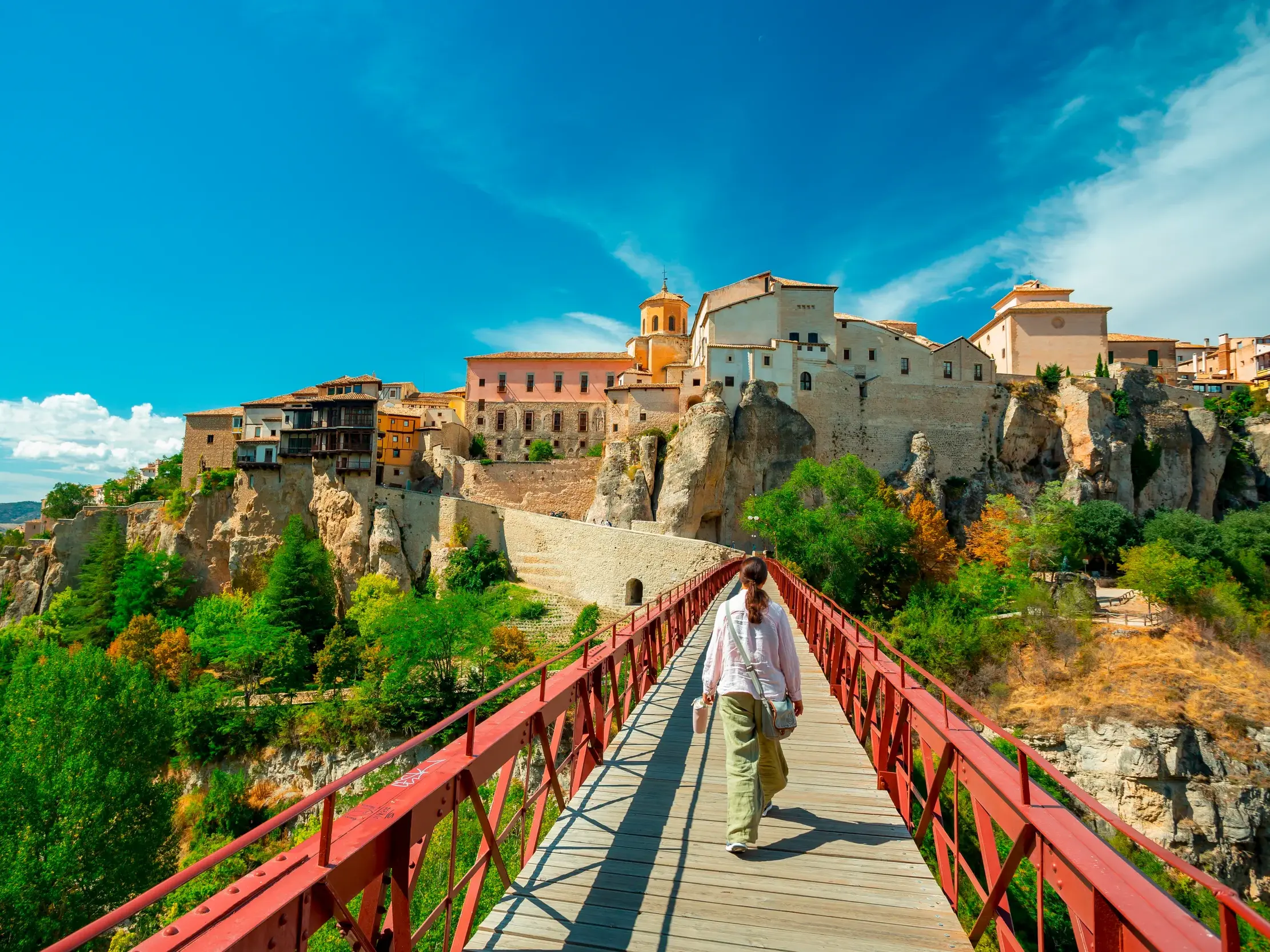 Eine Reisende überquert eine rote Fußgängerbrücke zur Altstadt von Cuenca, die über der Schlucht des Flusses Huécar thront.