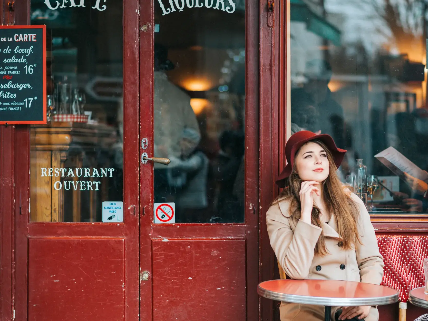 Eine Frau mit rotem Hut sitzt an einem Tisch vor einem Restaurant in Paris und genießt nachdenklich die Atmosphäre der Stadt.