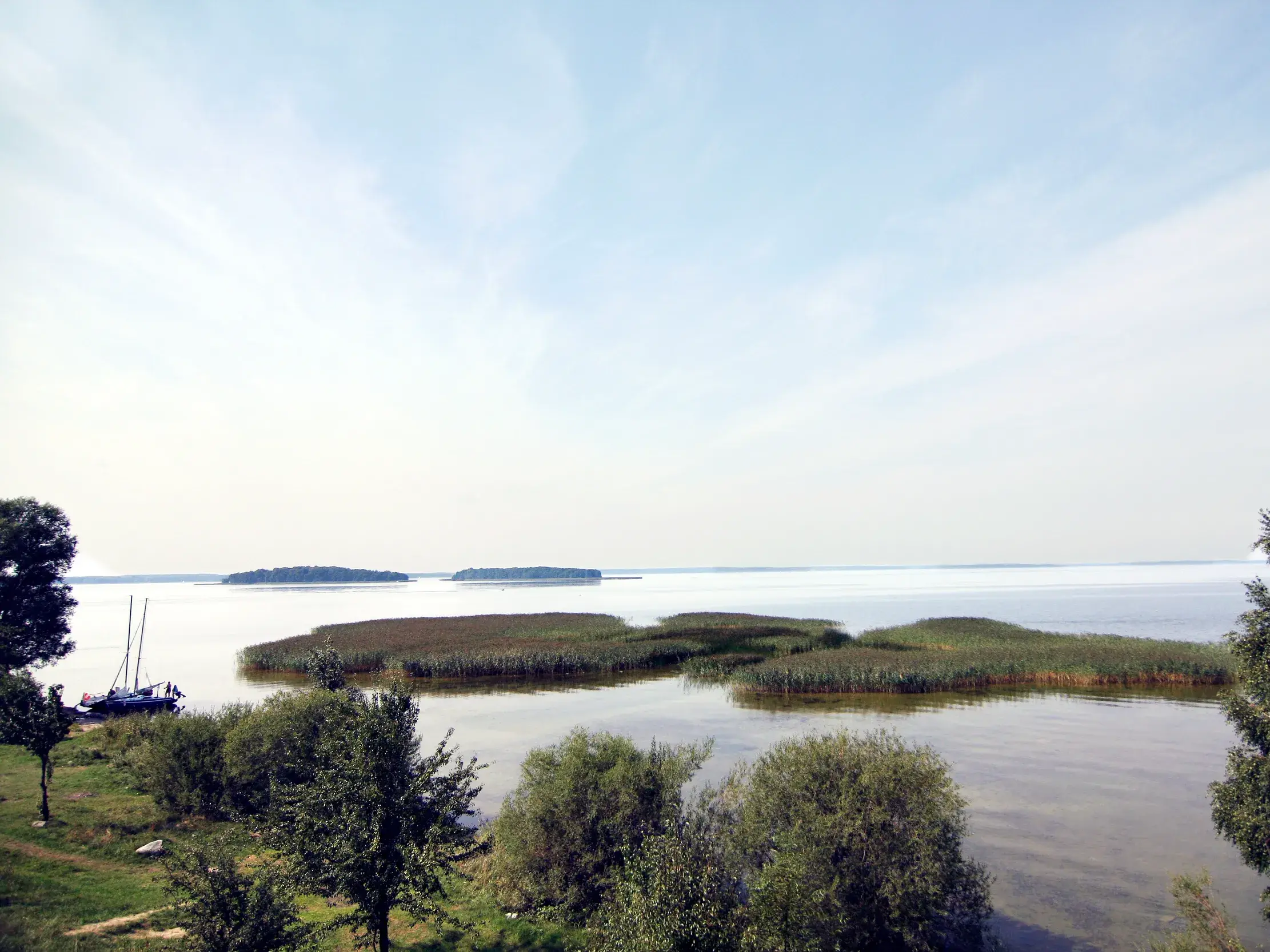 Weite Sicht über die Masurische Seenplatte mit Schilfinseln, bewaldeten Eilanden und einem Segelboot am Ufer.