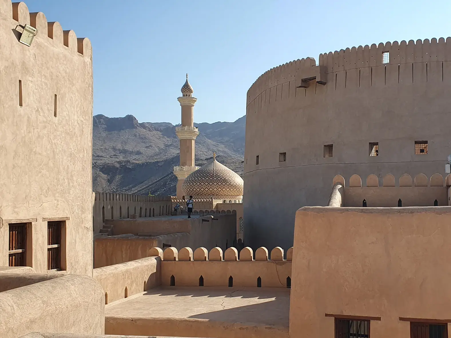 Sonniger Ausblick vom Nizwa Fort auf die verzierte Moschee-Kuppel vor der Kulisse des Hadschar-Gebirges im Oman.