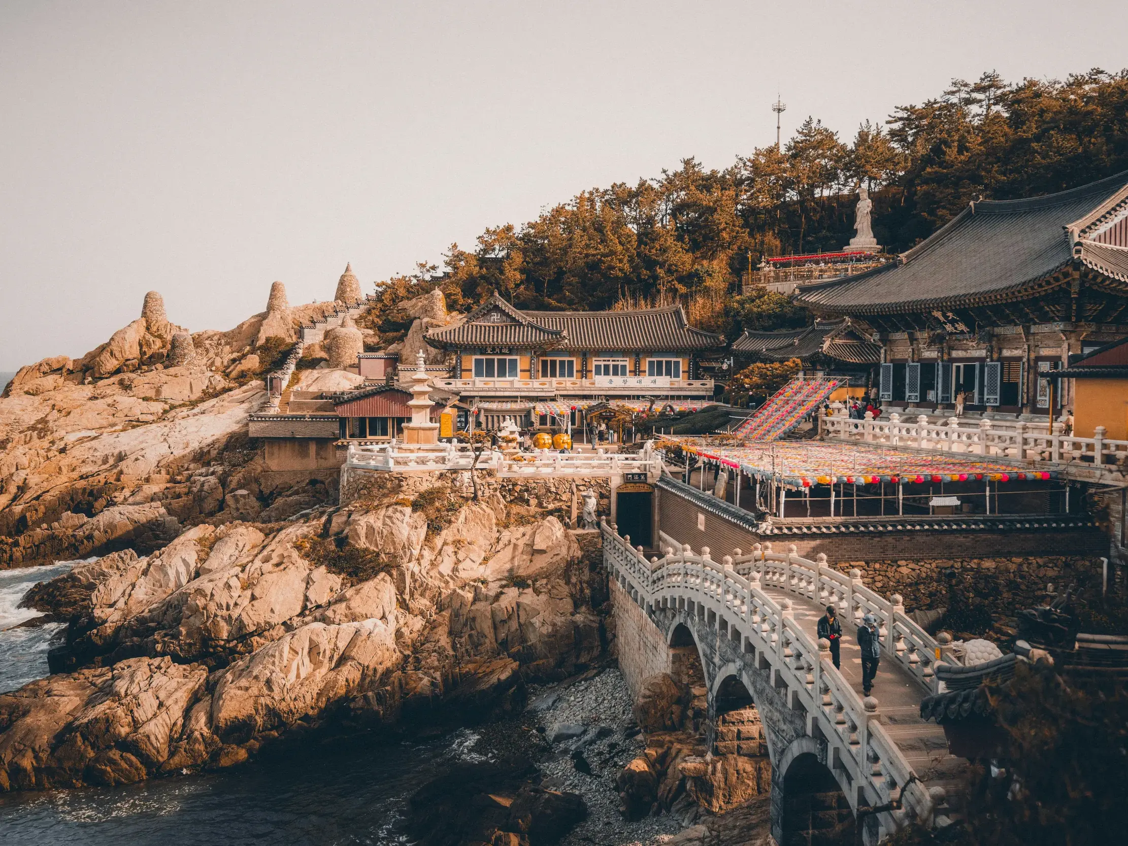Zwei Wanderer überqueren eine Steinbrücke zum Haedong Yonggungsa-Tempel, der an der felsigen Küste von Busan liegt.