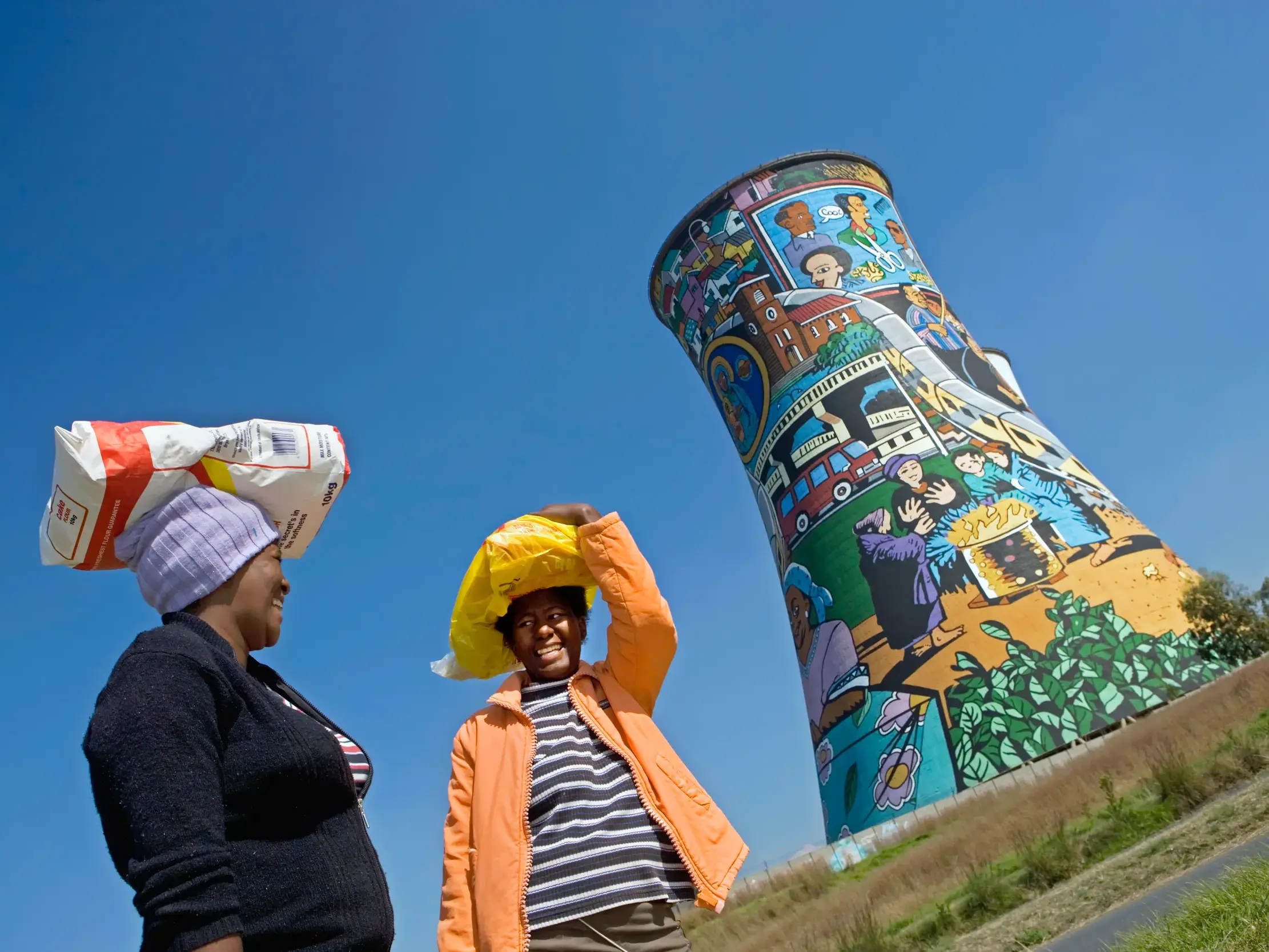 Zwei Frauen aus Soweto mit Lasten auf dem Kopf vor den farbenfroh bemalten Orlando Towers unter blauem Himmel in Johannesburg.