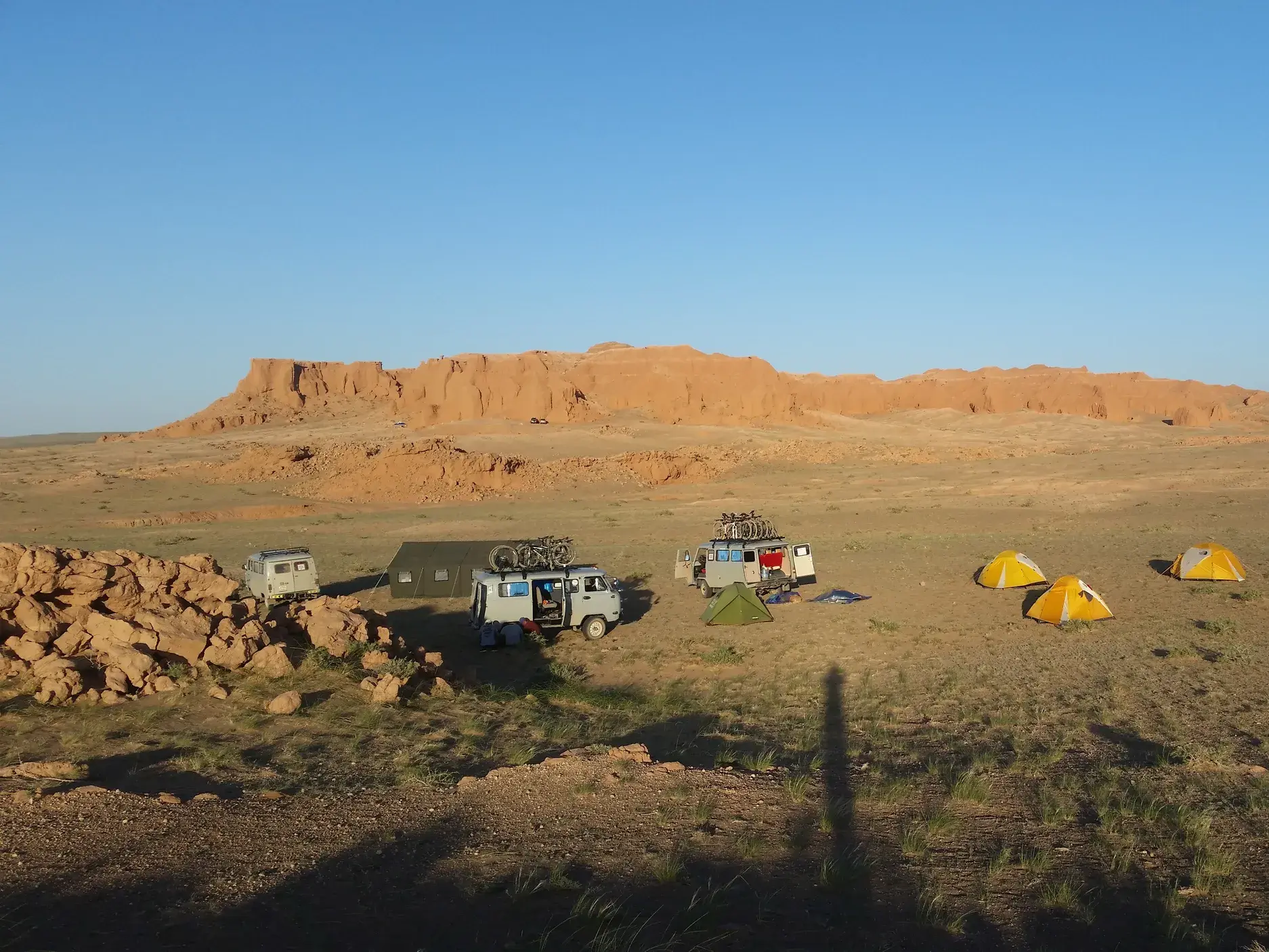 Zeltlager einer Radreise mit Begleitfahrzeugen in der mongolischen Gobi-Wüste vor den rot leuchtenden Klippen von Bayanzag.