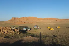 Zeltlager einer Radreise mit Begleitfahrzeugen in der mongolischen Gobi-Wüste vor den rot leuchtenden Klippen von Bayanzag.