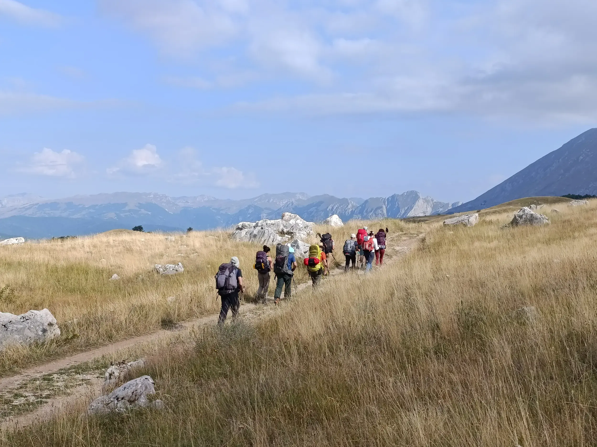 Eine Trekking-Gruppe wandert auf einem schmalen Pfad durch eine weite, sonnige Graslandschaft mit einer Bergkette am Horizont.