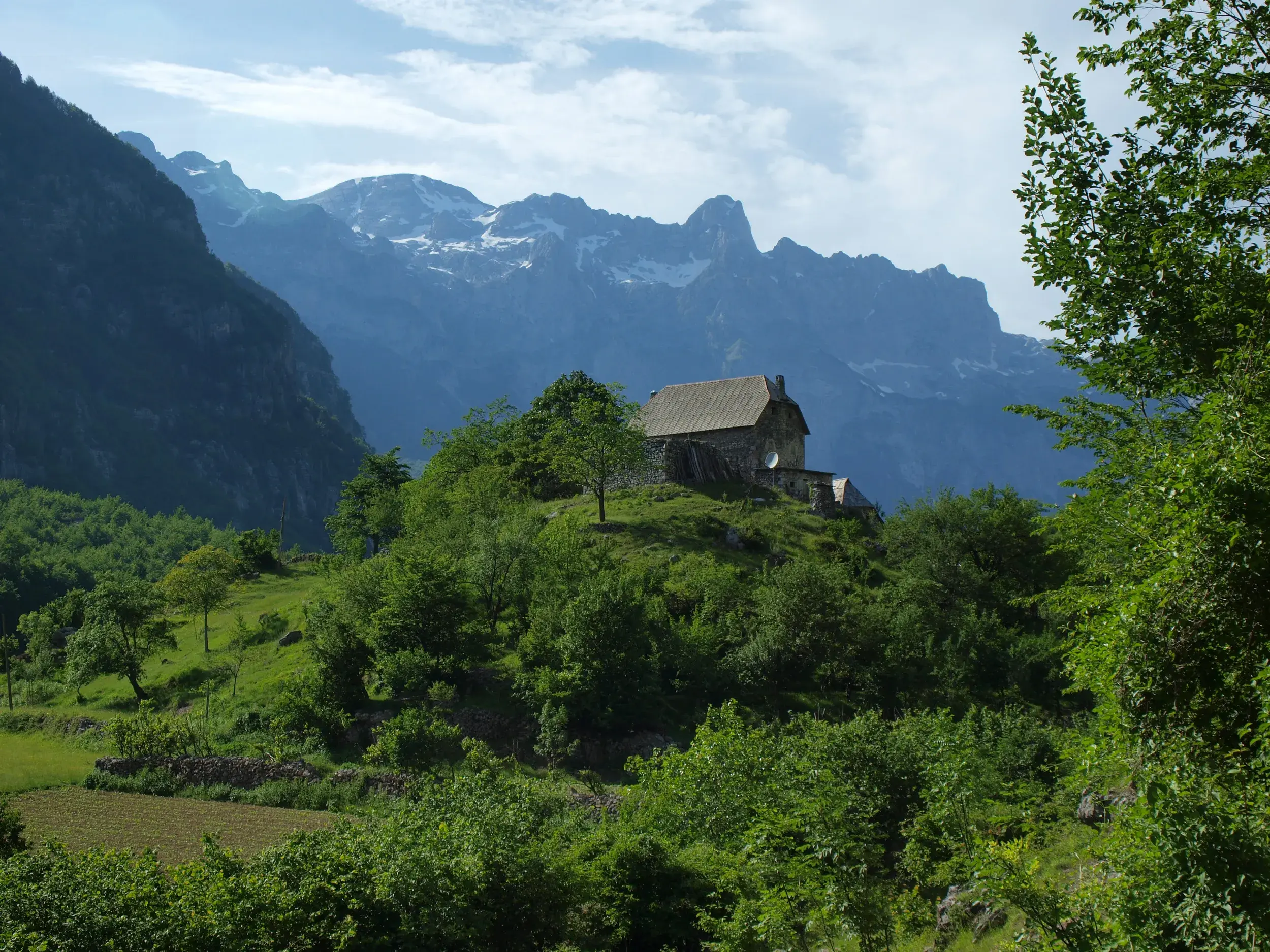 Abgelegenes Steinhaus auf einem bewaldeten Hügel vor der majestätischen Bergkulisse des Valbona-Nationalparks in Albanien.