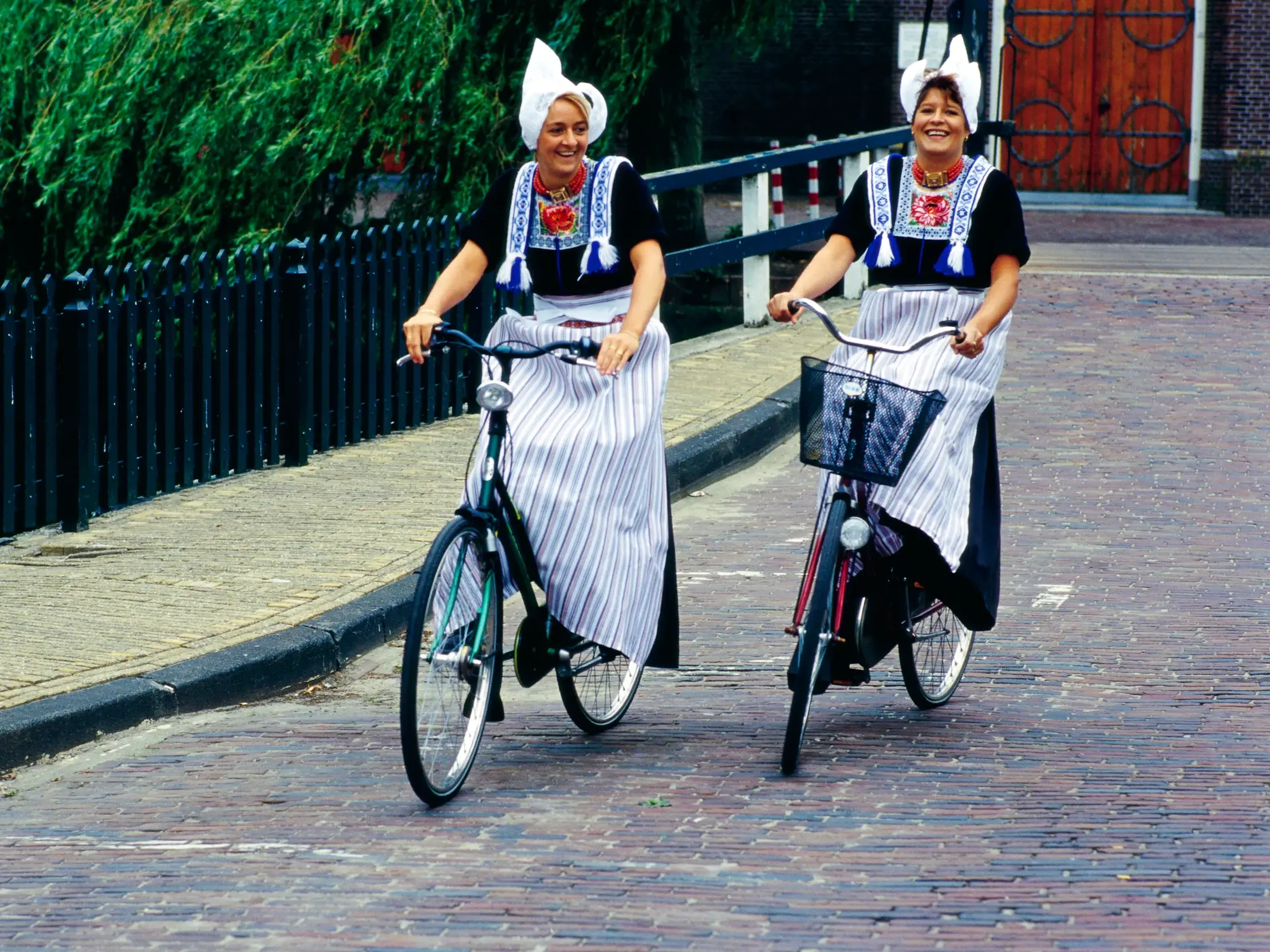 Zwei Frauen in holländischer Tracht erleben die Hollandromantik bei einer Fahrradfahrt auf einer Kopfsteinpflasterstraße.