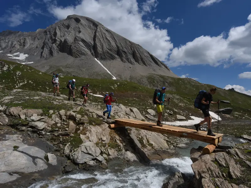 Eine Hauser-Wandergruppe überquert auf einer Alpenüberquerung eine Holzbrücke vor einem imposanten Alpengipfel.