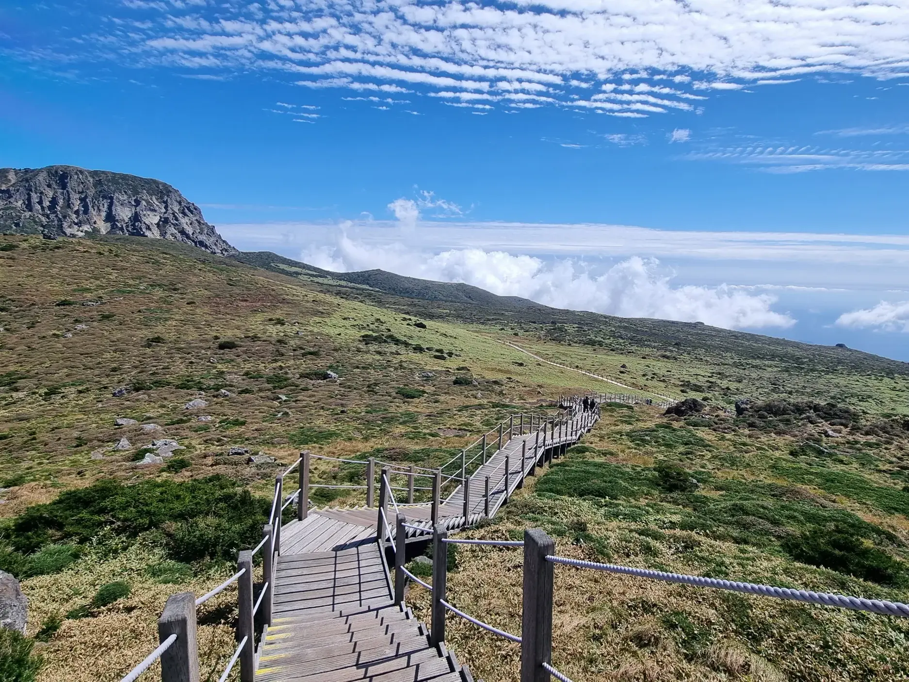 Wanderer auf einem Holzsteg des Yeongsil-Trails mit Blick auf die weite, vulkanische Landschaft des Hallasan in Südkorea.
