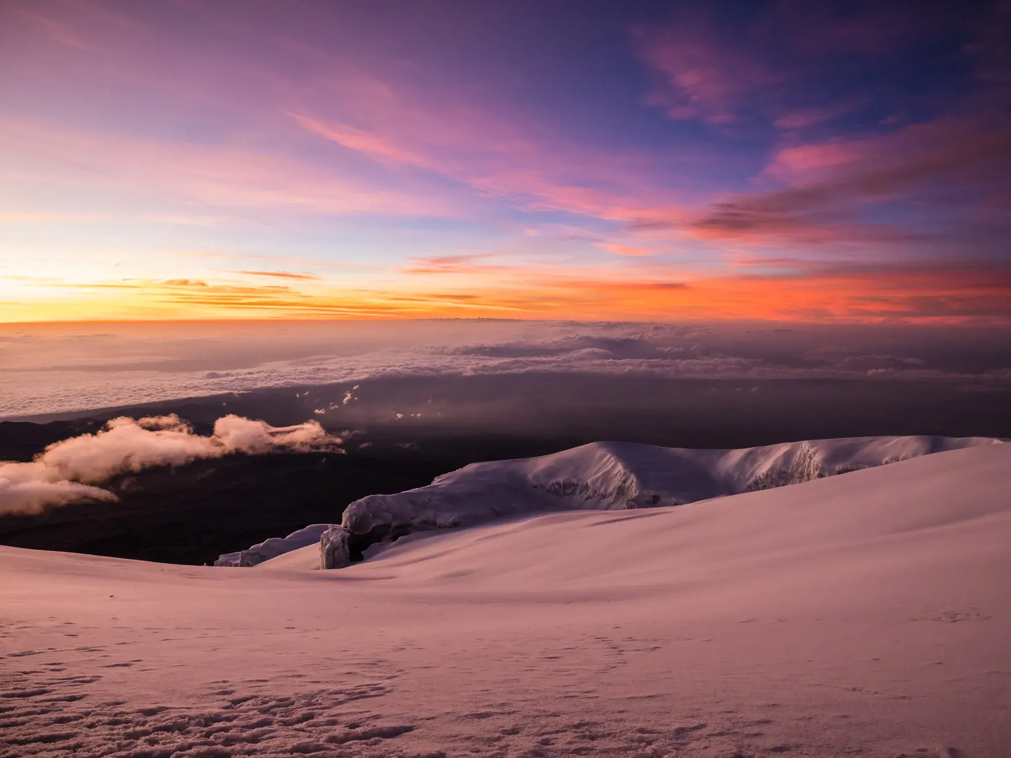 Sonnenaufgang am Kilimanjaro. Die schneebedeckte Gipfelregion leuchtet in rosa und orangen Farben über einem dichten Wolkenmeer.