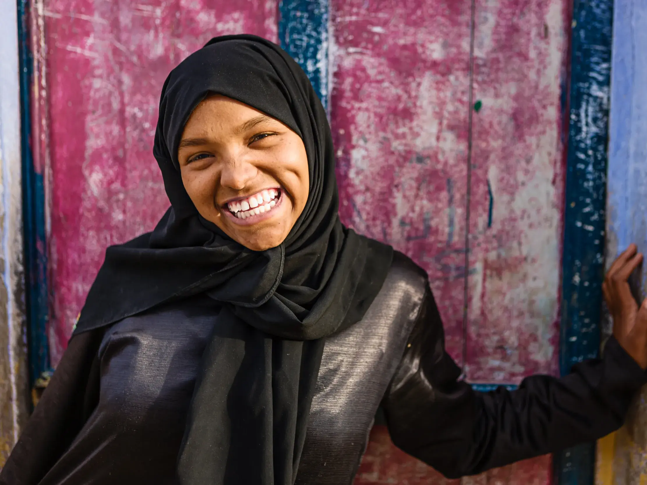 Eine junge ägyptische Frau mit schwarzem Hijab lacht herzlich und blickt in die Kamera vor einer bunten Wand.
