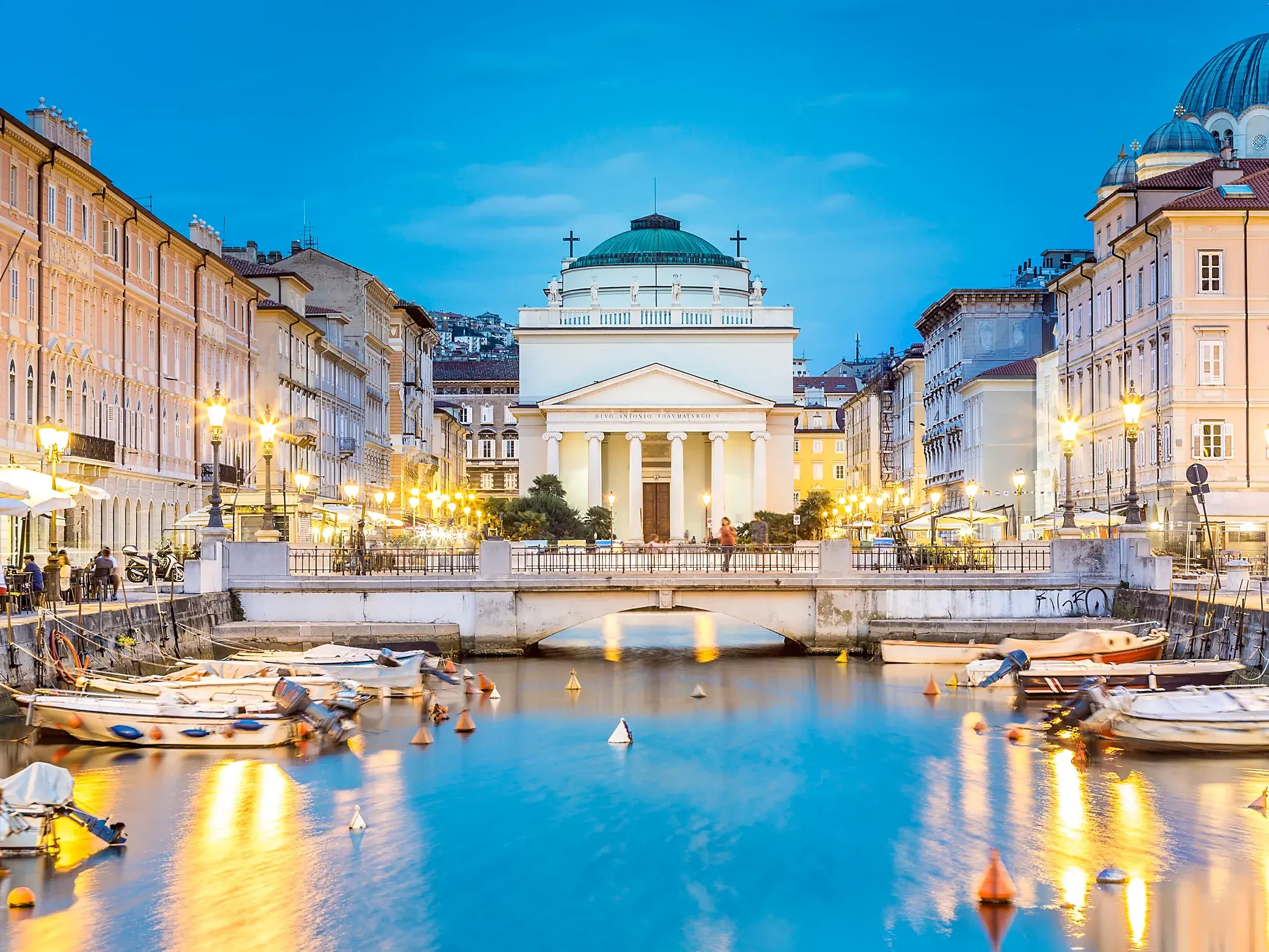 Die Kirche Sant'Antonio Taumaturgo am Ende des Canal Grande in Triest bei blauer Stunde, mit Booten und beleuchteten Fassaden.