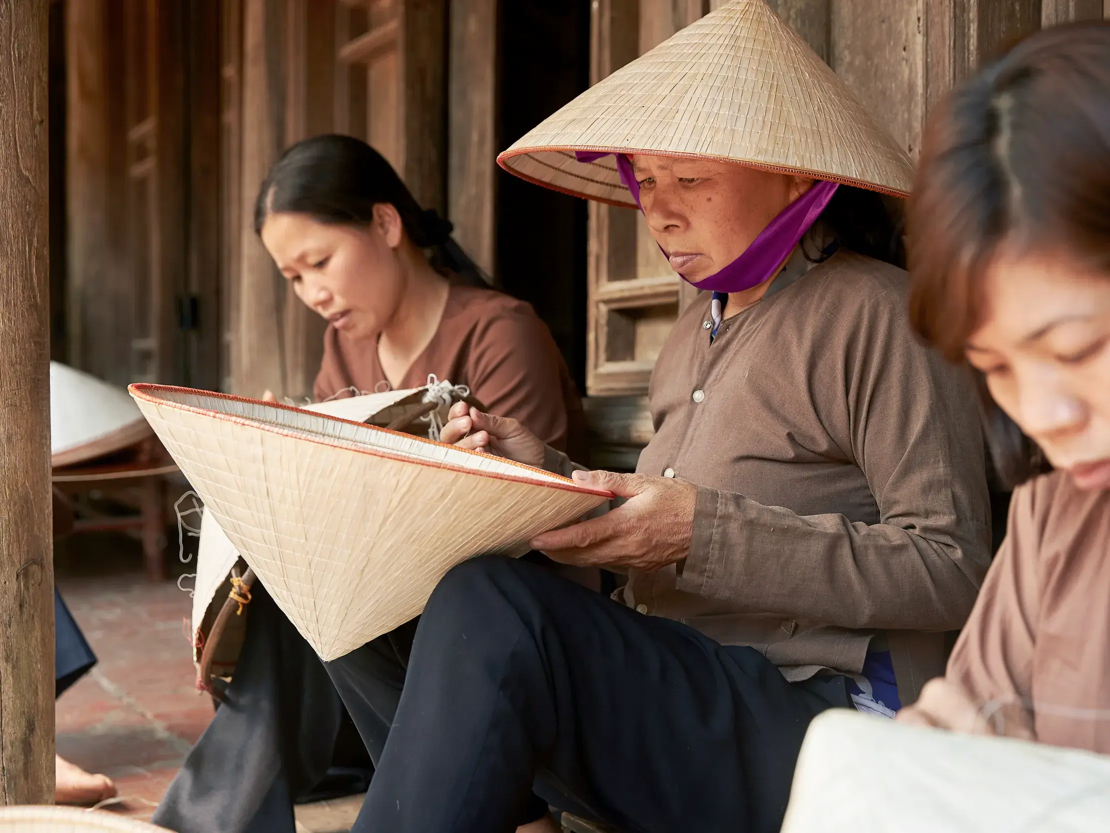 Drei vietnamesische Frauen fertigen in konzentrierter Handarbeit traditionelle Kegelhüte, die Nón Lá, vor einem Holzhaus.