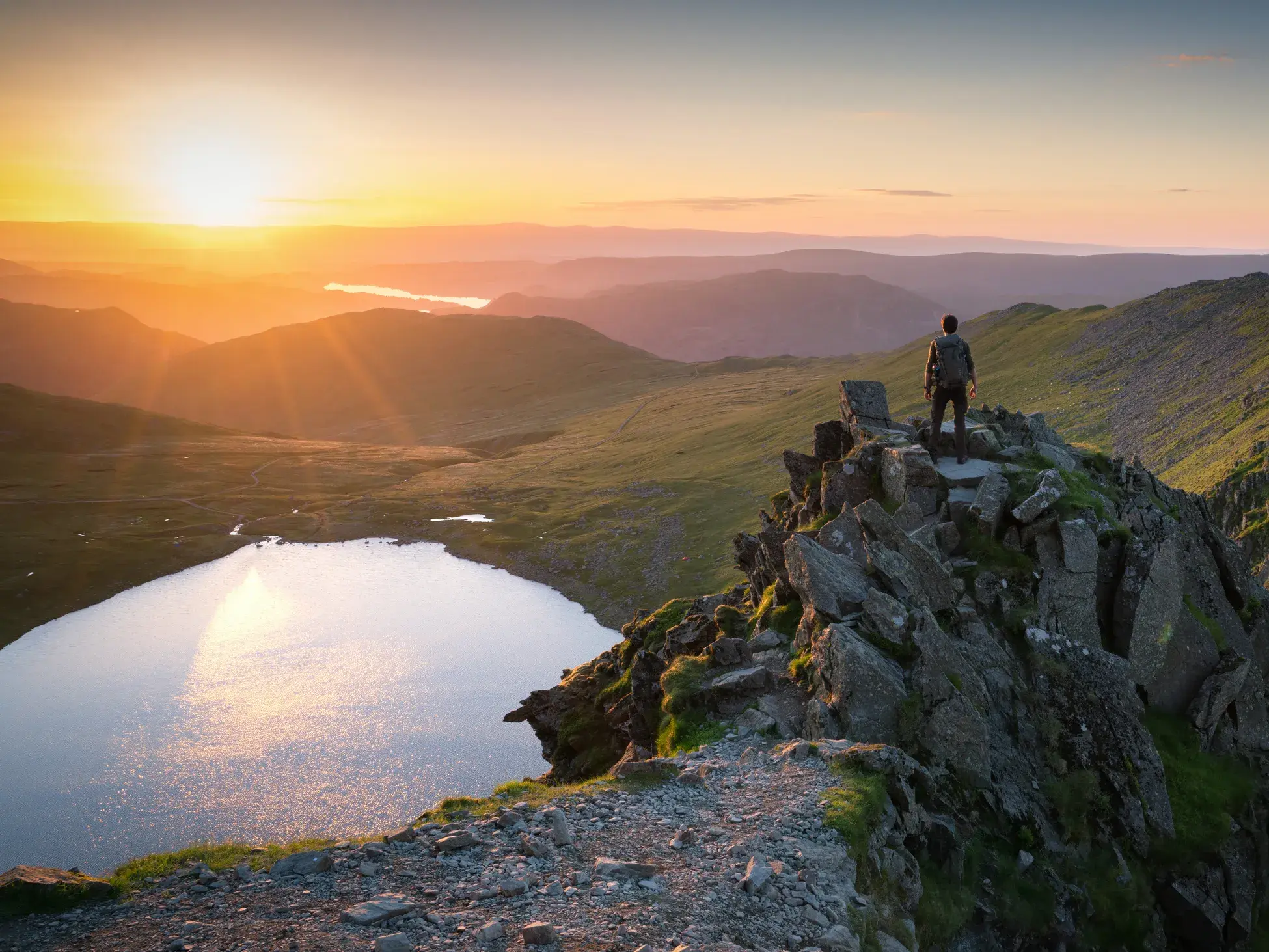 Ein Trekker genießt den Sonnenuntergang von einem Gipfel mit Blick über die Seen- und Berglandschaft des englischen Lake District.