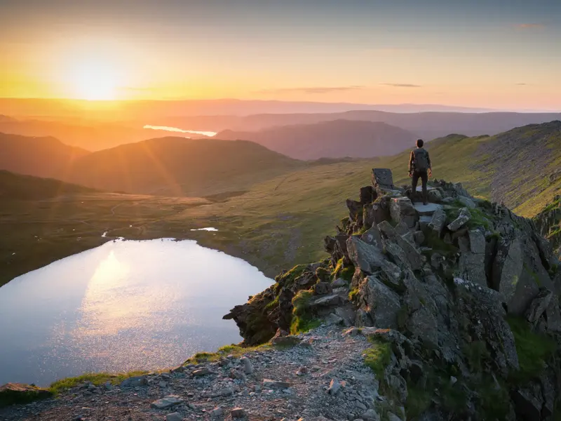 Ein Trekker genießt den Sonnenuntergang von einem Gipfel mit Blick über die Seen- und Berglandschaft des englischen Lake District.