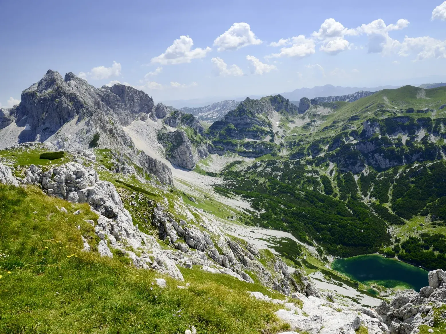 Blick von einem grasbewachsenen Berghang auf die Karstgipfel und einen tiefblauen Bergsee im Durmitor-Nationalpark.