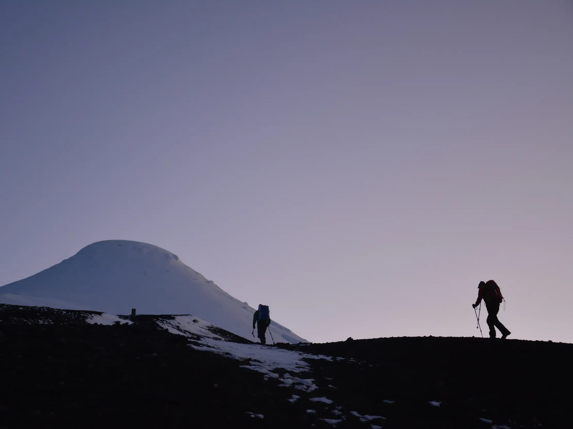 Zwei Trekker als Silhouetten auf einem Berggrat in der Dämmerung vor dem schneebedeckten Vulkankegel des Osorno in Chile.