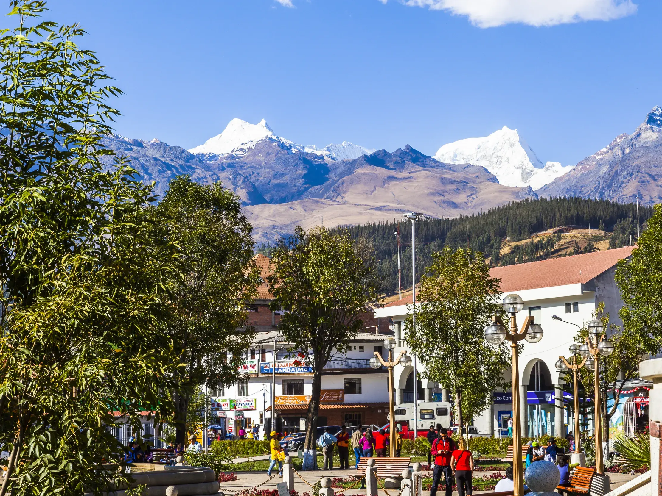 Belebter Stadtplatz in Huaraz mit grünen Bäumen. Im Hintergrund erheben sich die schneebedeckten Gipfel der Cordillera Blanca.