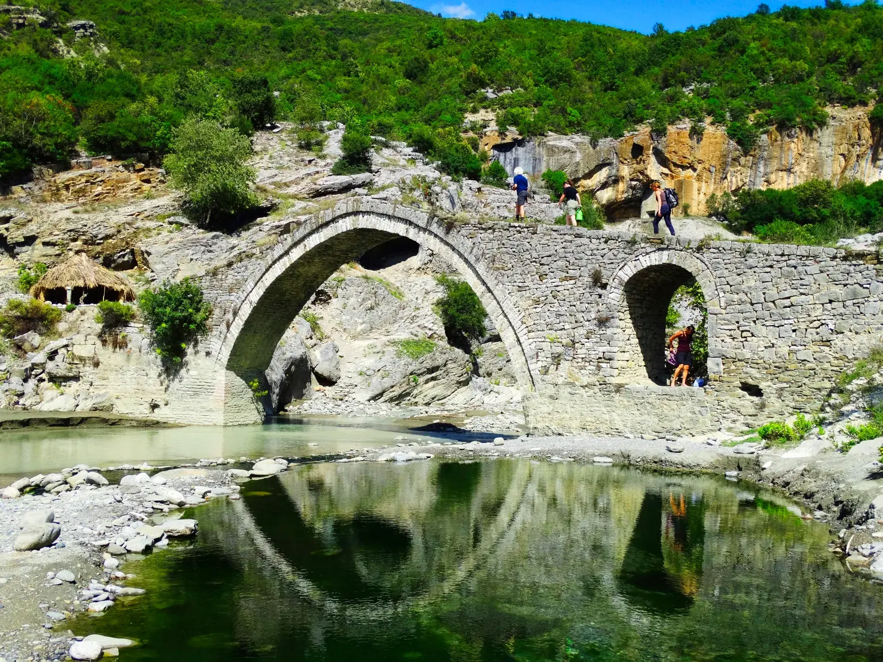 Wandergruppe bei der Erkundung der alten Bogenbrücke von Benja aus osmanischer Zeit in der grünen Berglandschaft Albaniens.