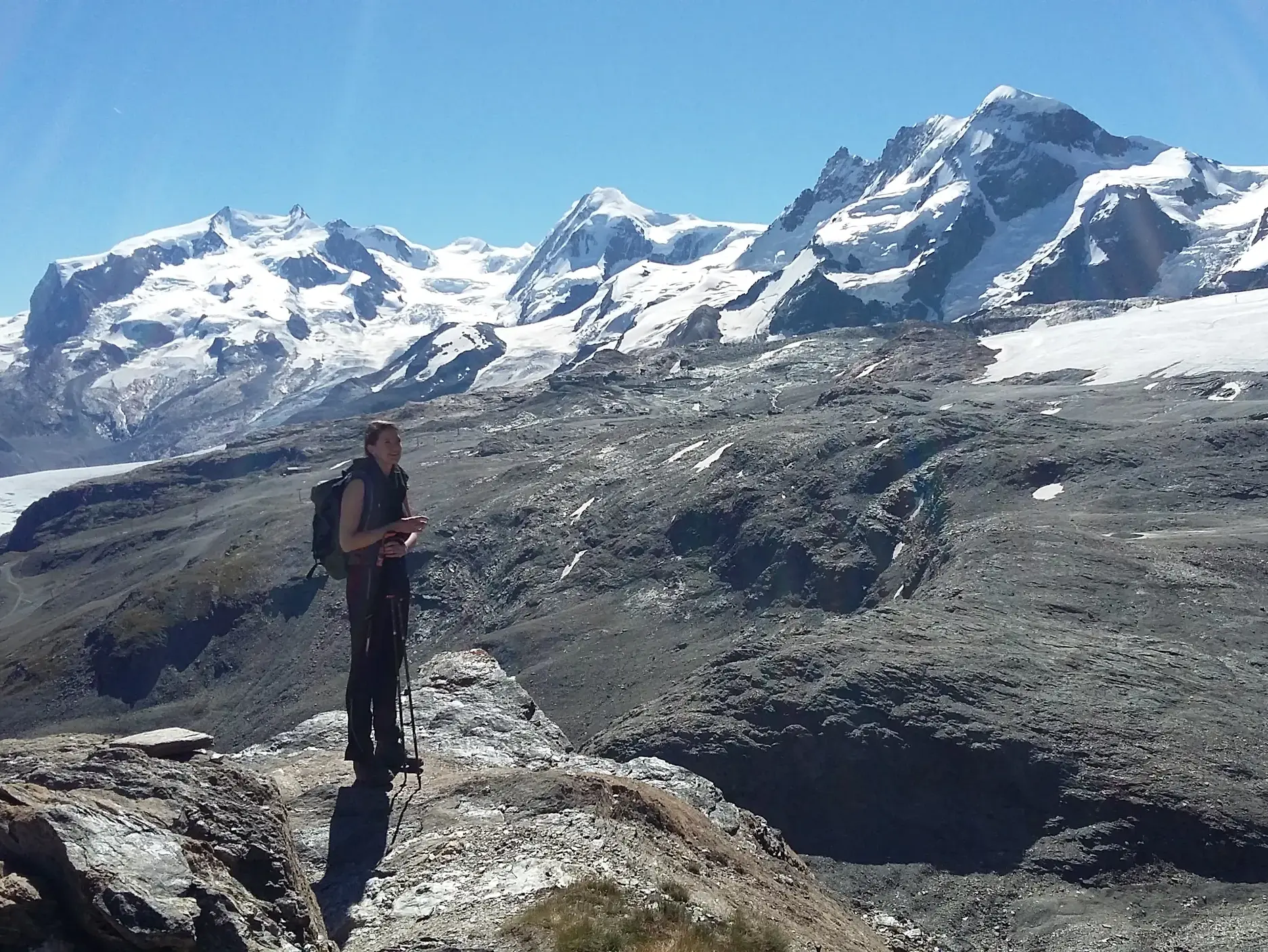Eine Wanderin mit Rucksack orientiert sich auf einem Höhenweg vor dem Panorama der Walliser Gletscher in der Nähe von Zermatt.