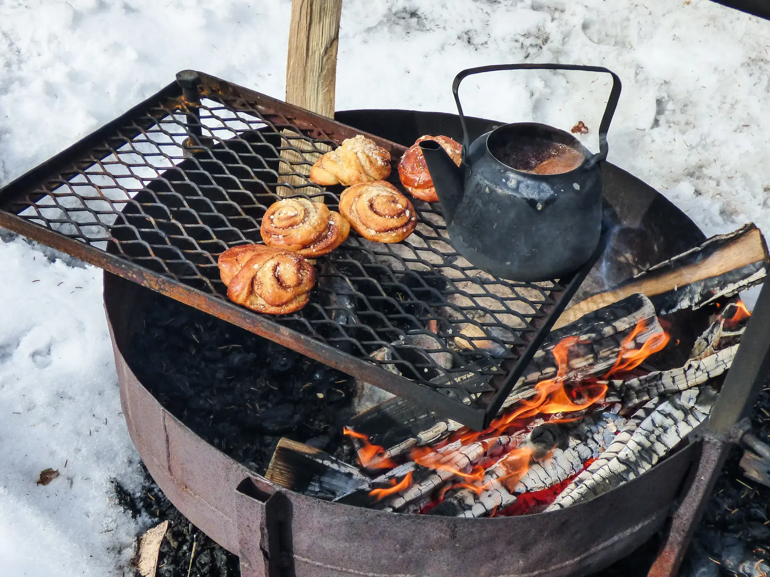 Zimtschnecken und eine schwarze Kaffeekanne wärmen auf einem Grillrost über einem Lagerfeuer in der finnischen Schneelandschaft.