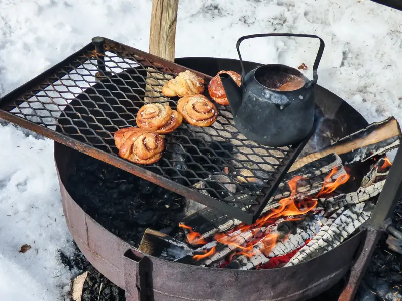 Zimtschnecken und eine schwarze Kaffeekanne wärmen auf einem Grillrost über einem Lagerfeuer in der finnischen Schneelandschaft.