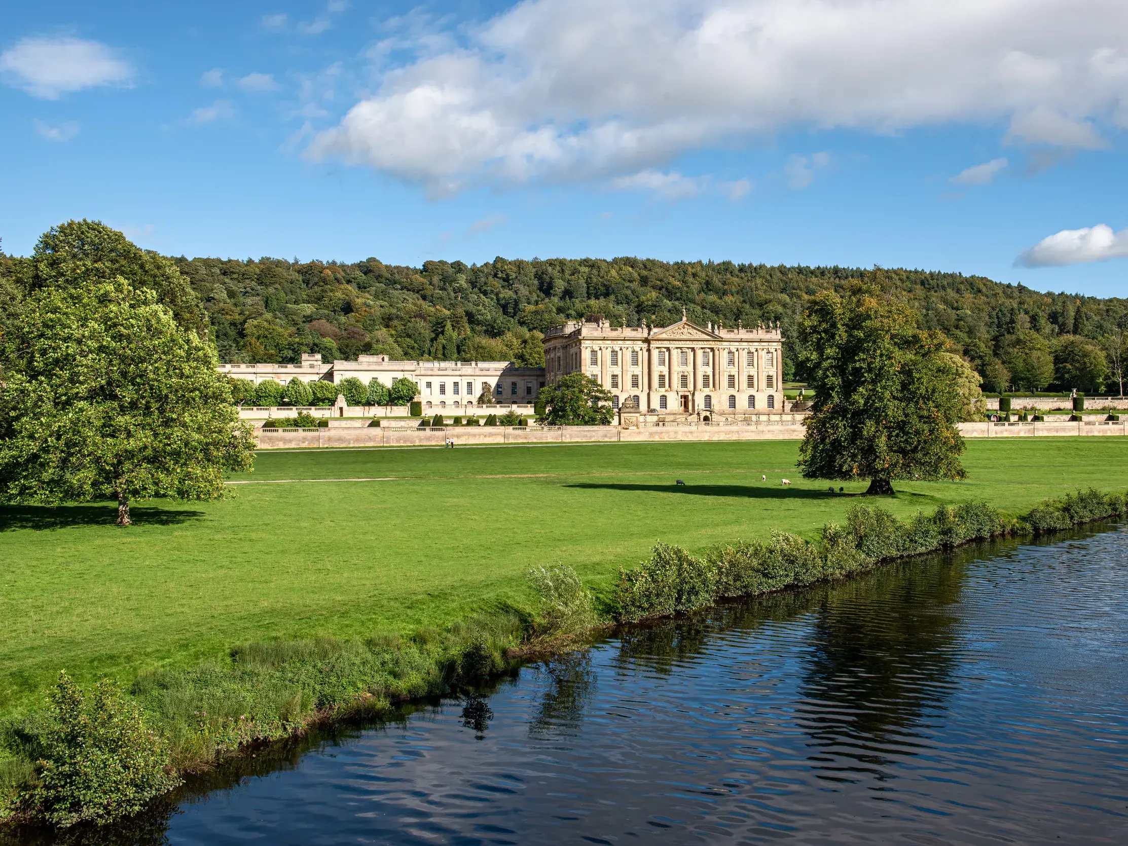 Das prächtige Herrenhaus Chatsworth House im Peak District, umgeben von Gärten und einem bewaldeten Hügel, am Ufer eines Flusses.