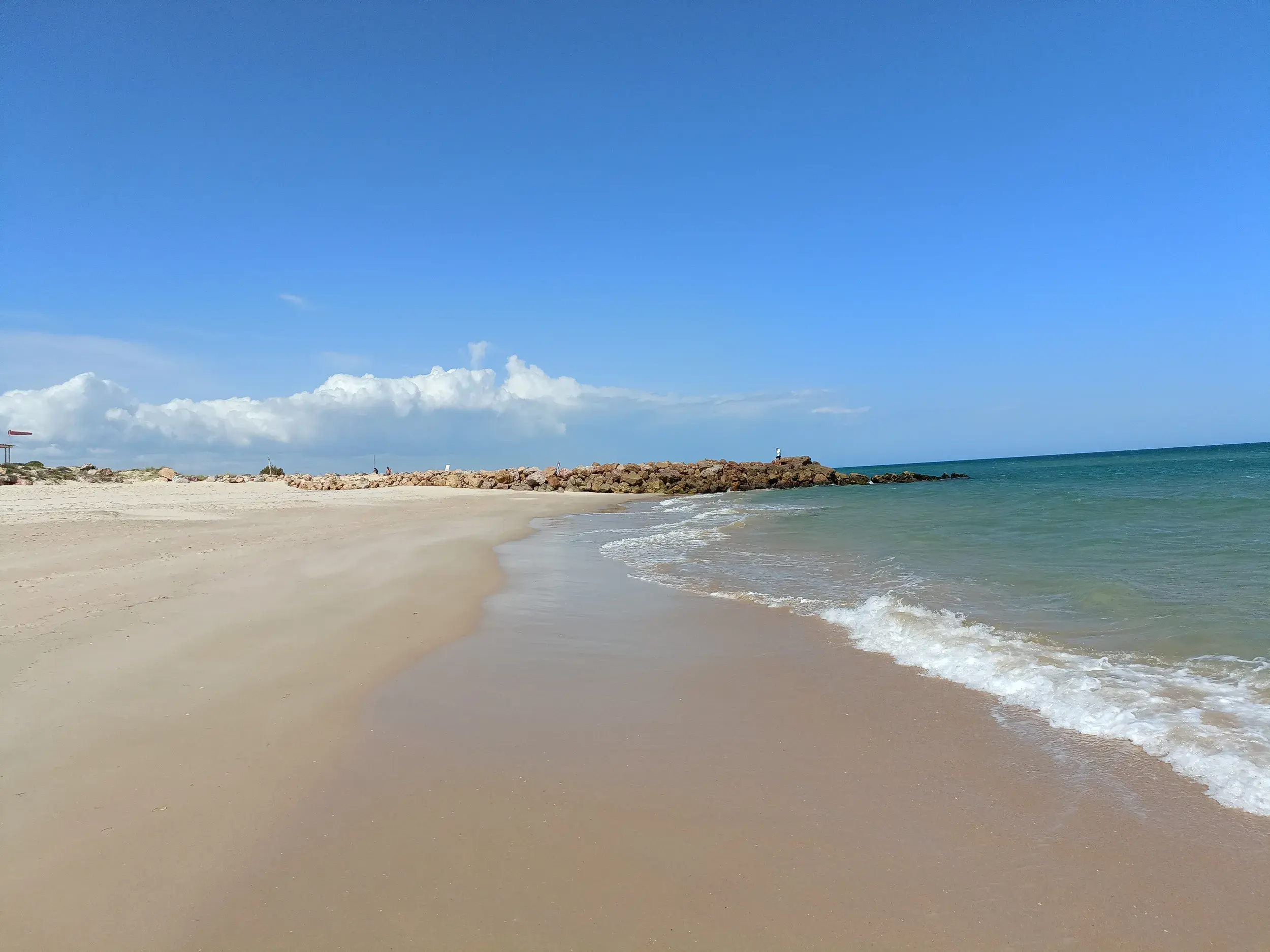 Wanderung am weiten Sandstrand des Naturparks Ria Formosa an der Algarve, mit Blick auf eine Steinmole und das Meer.