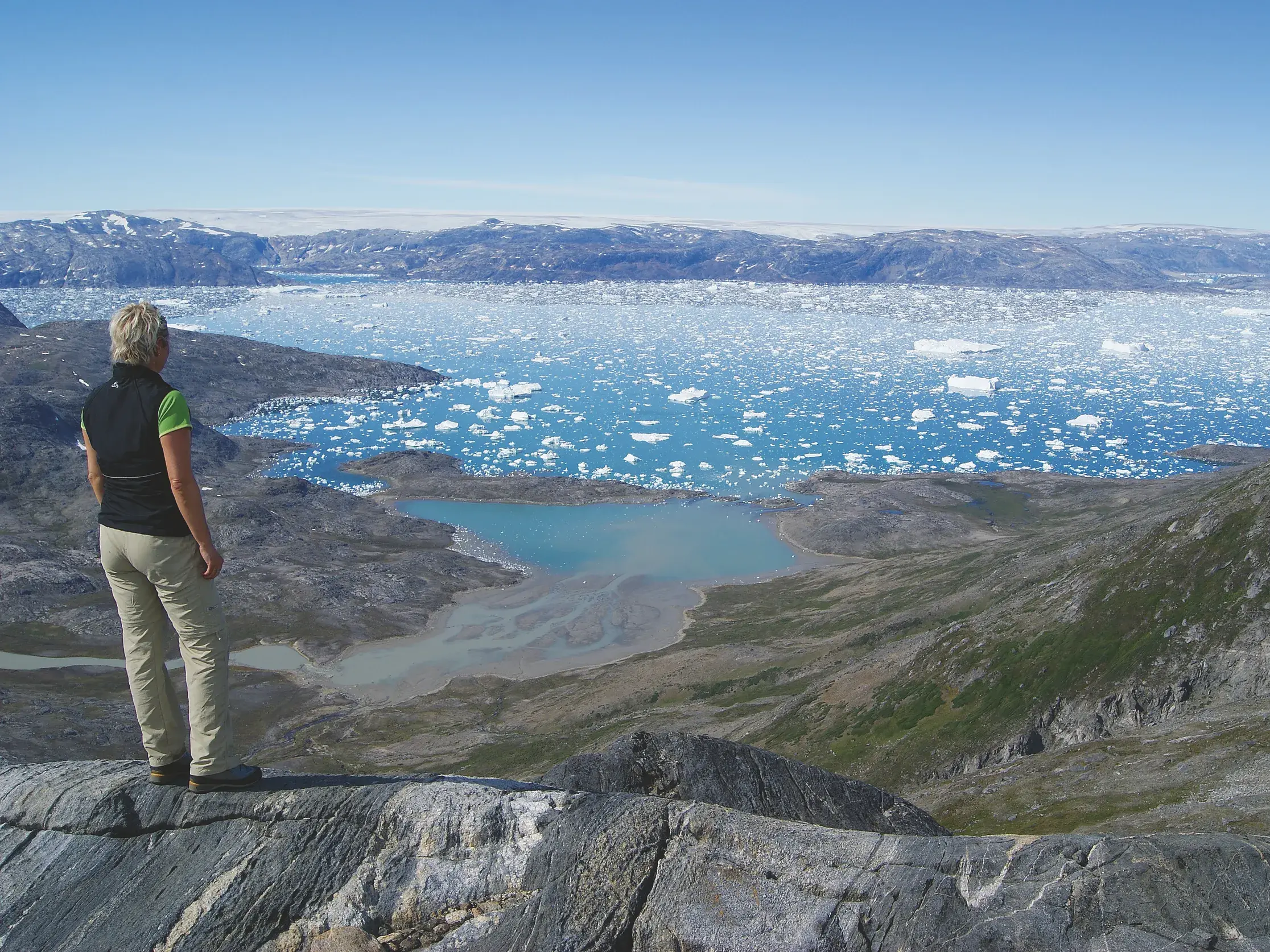 Eine Wanderin auf einem Felsen mit Panoramablick über den weiten Sermilik-Fjord voller Eisberge in Ostgrönland.