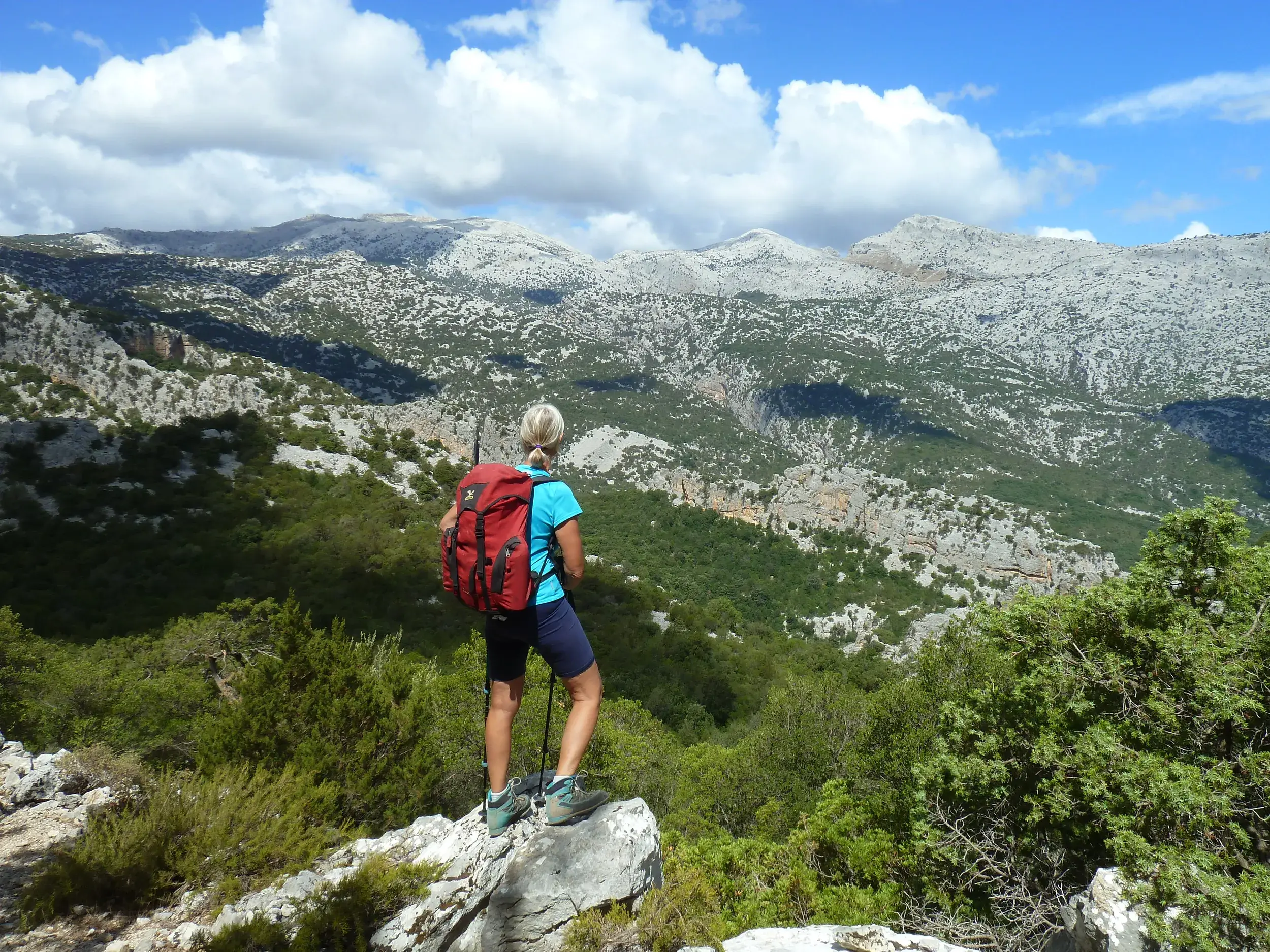 Eine Wanderin mit rotem Rucksack und Stöcken genießt den Ausblick auf die wilde Berglandschaft des Supramonte auf Sardinien.