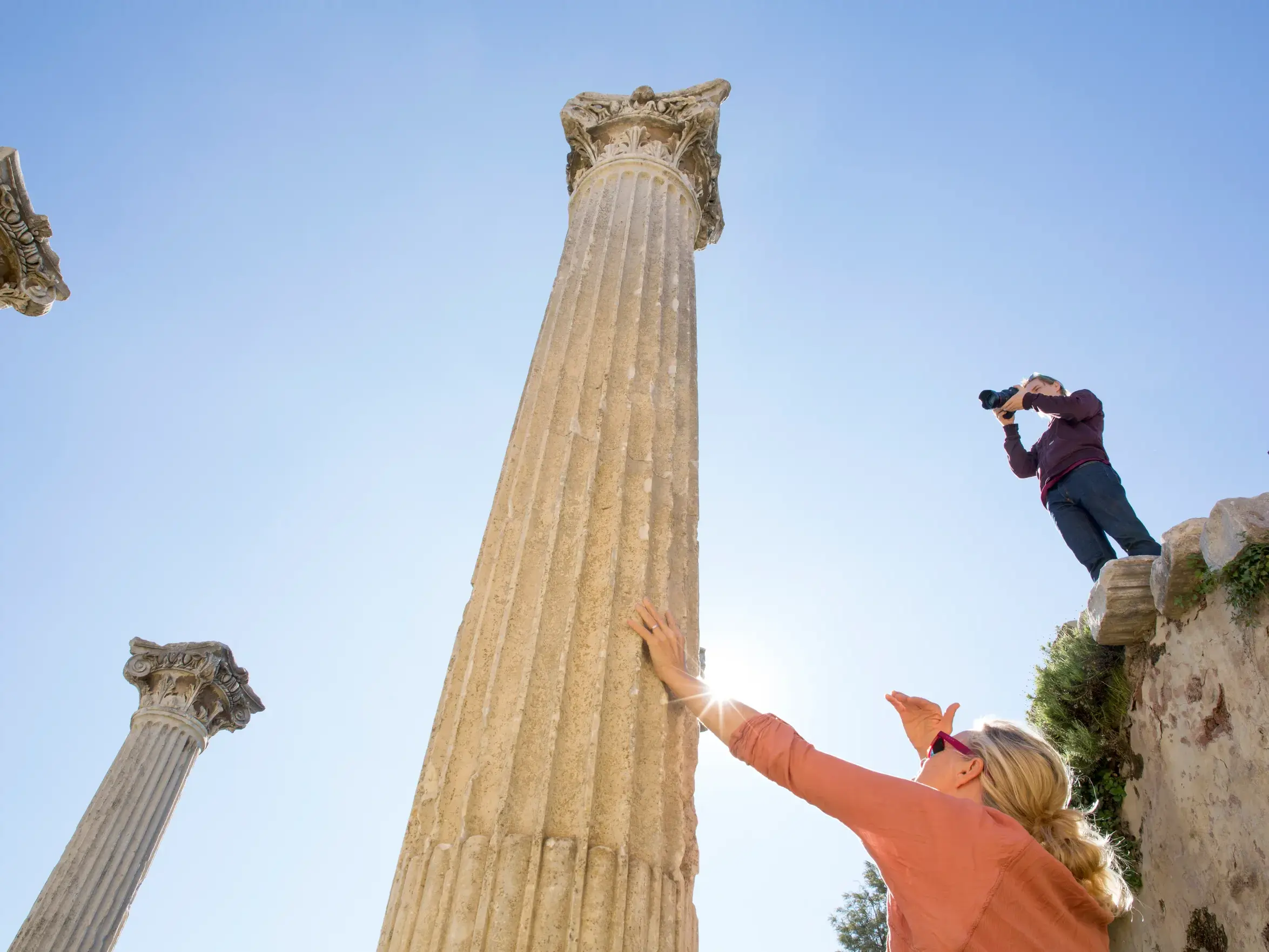 Frau berührt eine hohe antike Säule in den Ruinen von Pergamon, während ein Mann auf einer Mauer sie fotografiert.