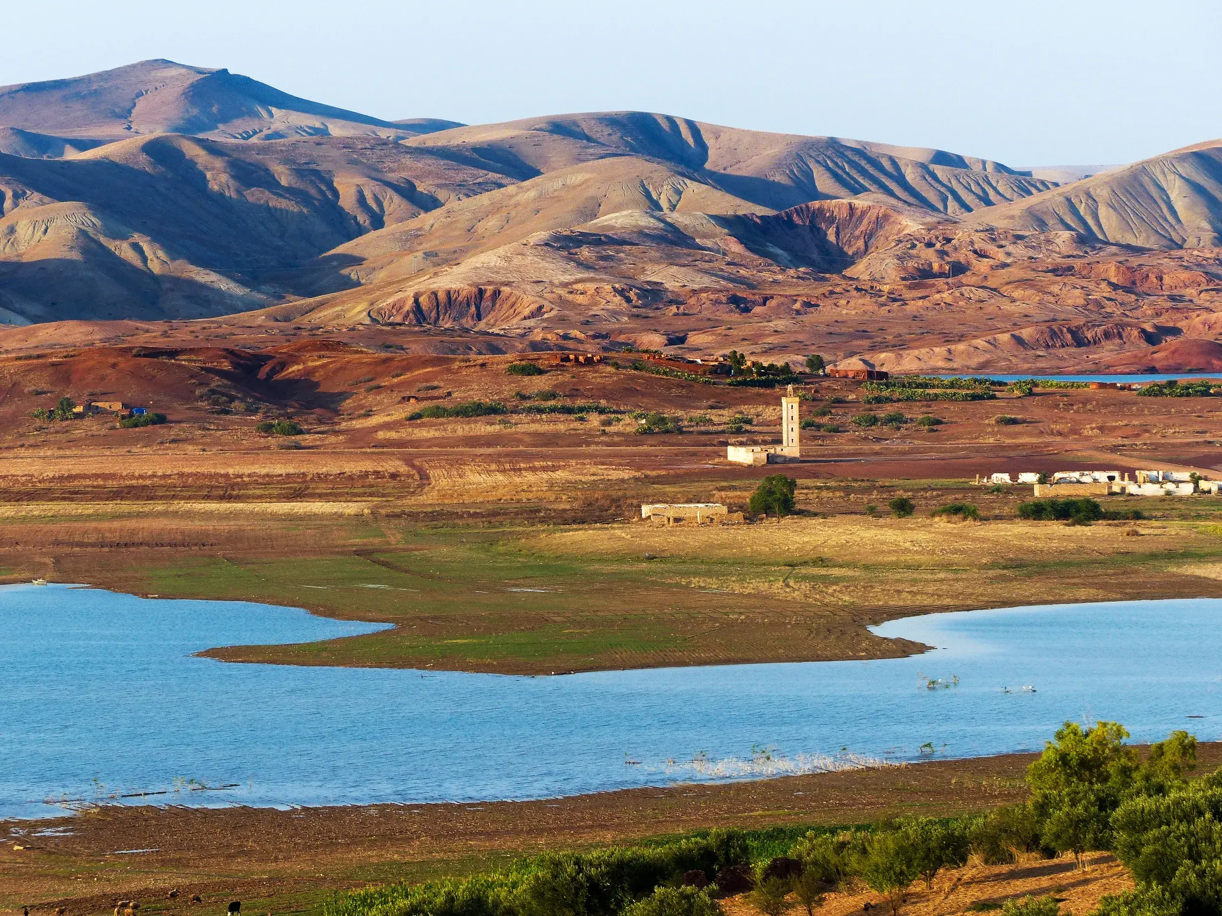 Weite, karge Landschaft des marokkanischen Atlasgebirges mit einem blauen Stausee und einem einsamen Minarett am Ufer.