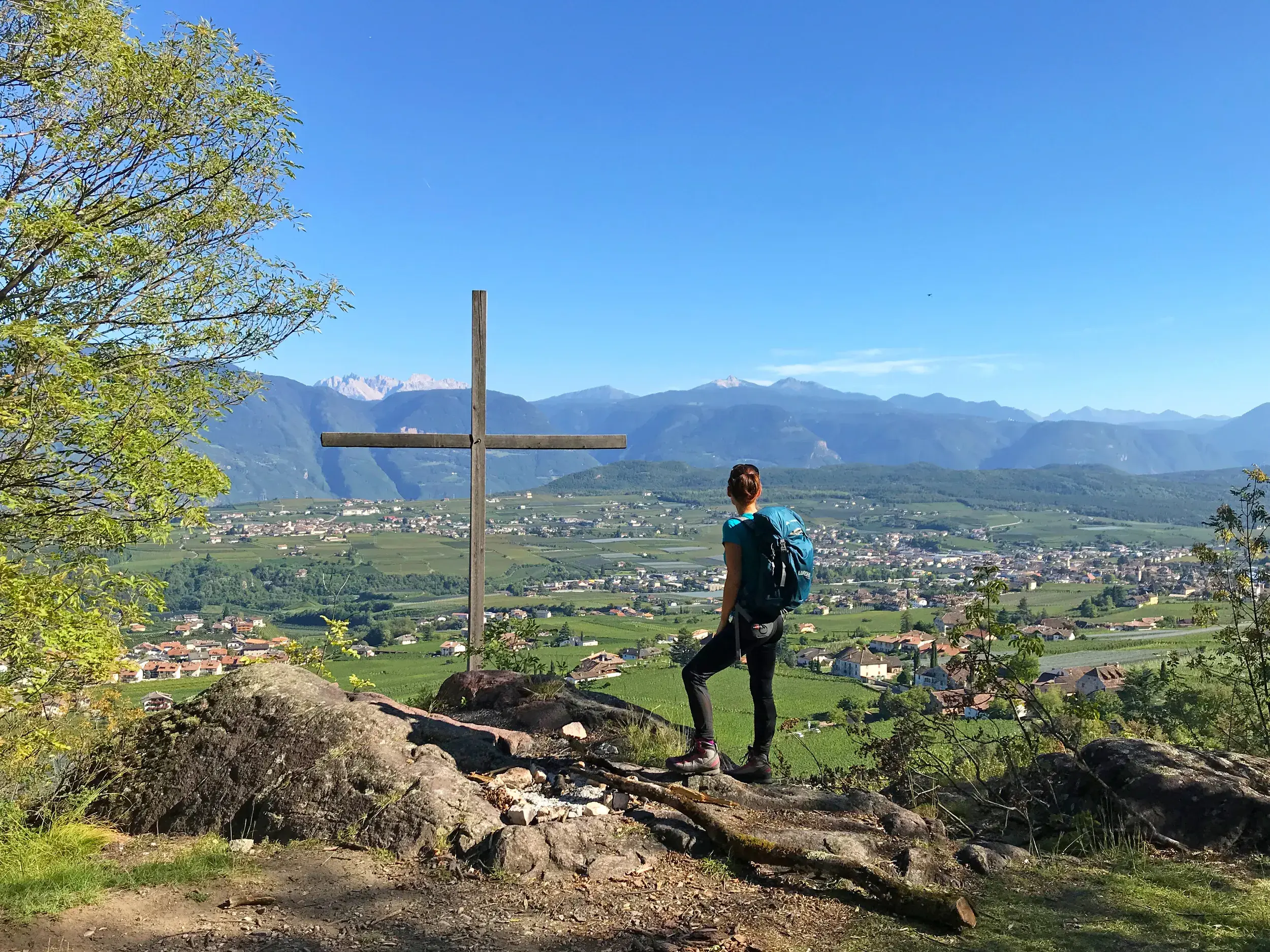 Wanderin mit Rucksack genießt an einem Holzkreuz die Aussicht über das sonnige Etschtal in Südtirol mit seinen Weinbergen.