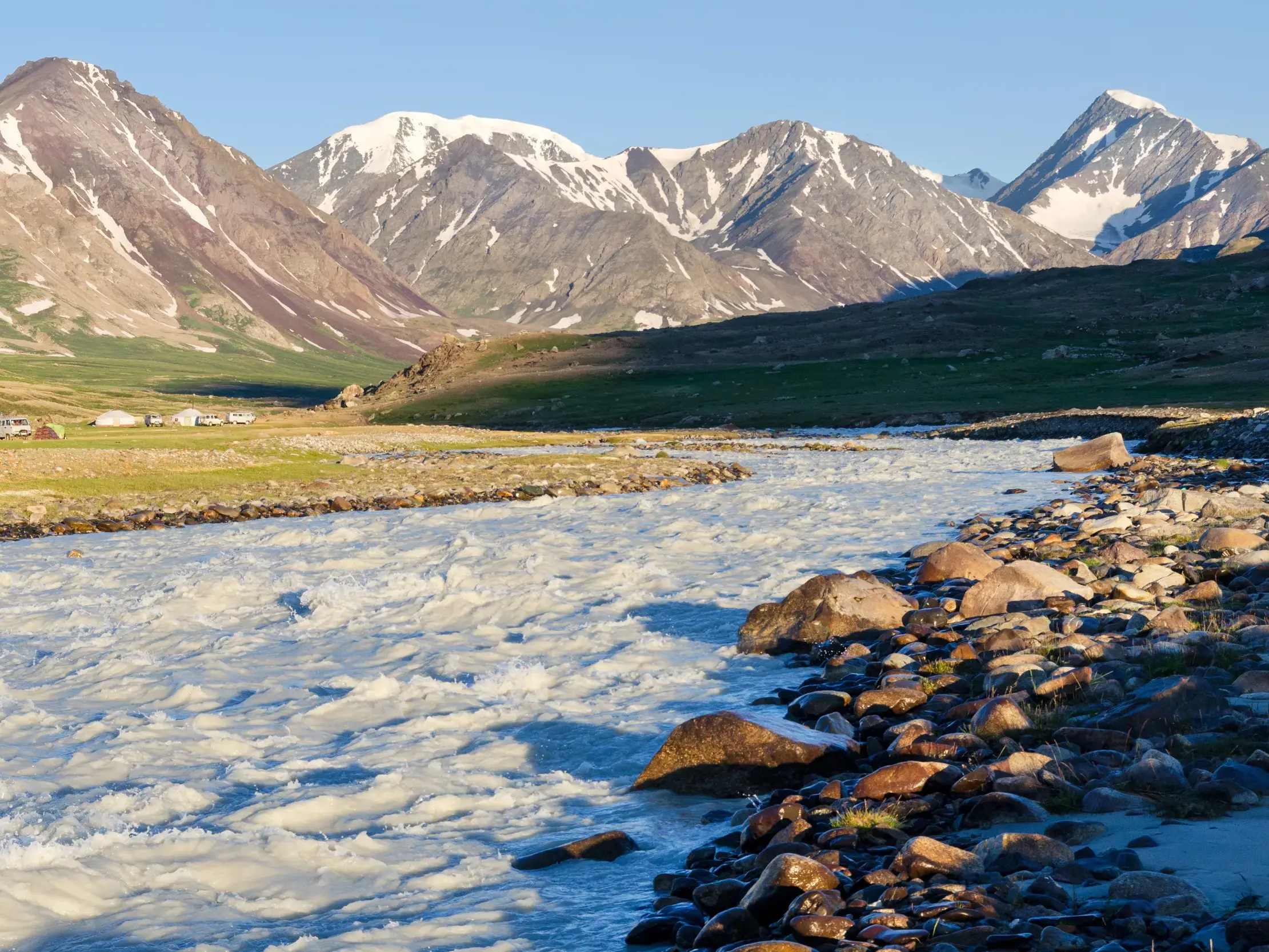 Zeltlager am reißenden Fluss Tsagaan Gol in der Mongolei, im Hintergrund die schneebedeckten Gipfel des Altai-Gebirges.